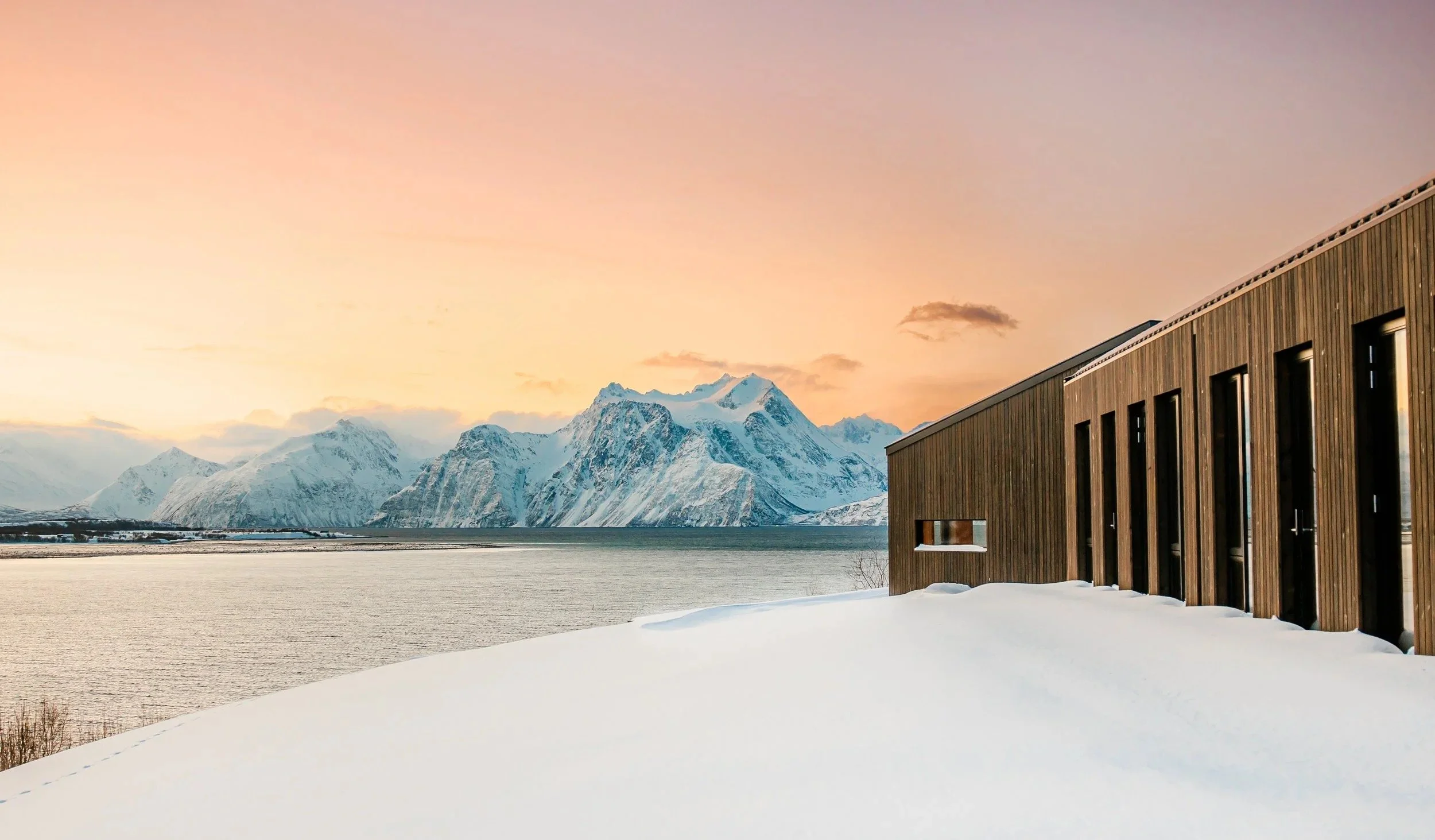 Modern wooden lodge in a snow-covered winter landscape overlooking a fjord and snowy mountains at sunset.