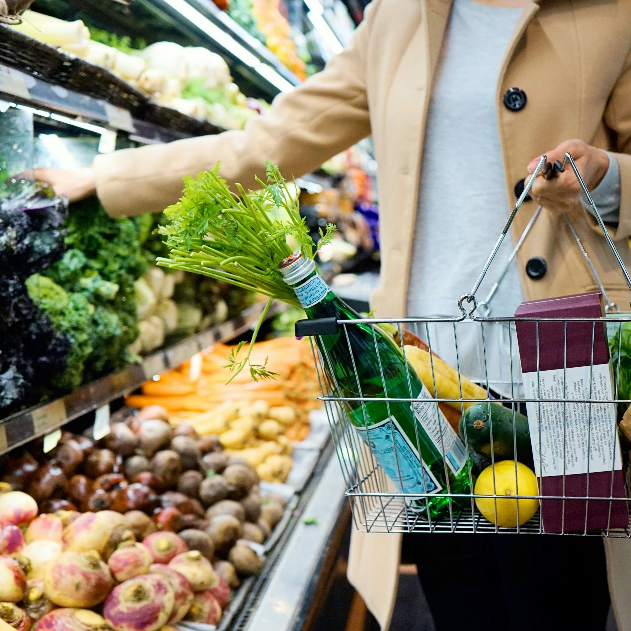 Einkaufswagen mit einer Wasserflasche, Zitrone, Gurke und einem Stück Papier im Supermarkt, im Hintergrund Obst- und Gemüsesortimente.