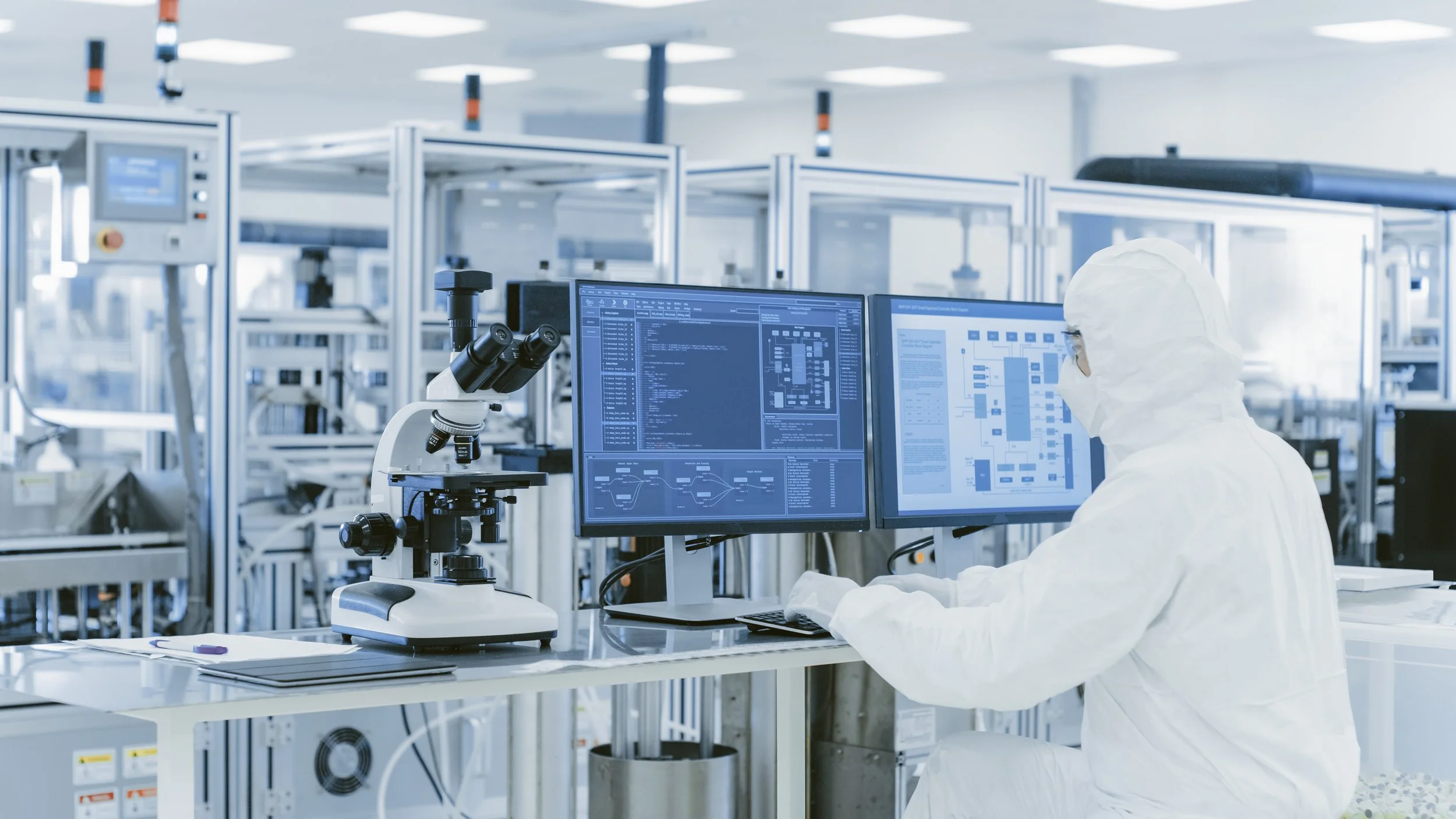 Ein Wissenschaftler in einem Reinraum arbeitet an einem Computer mit mehreren Monitoren, umgeben von technischen Geräten und Ausrüstung.
