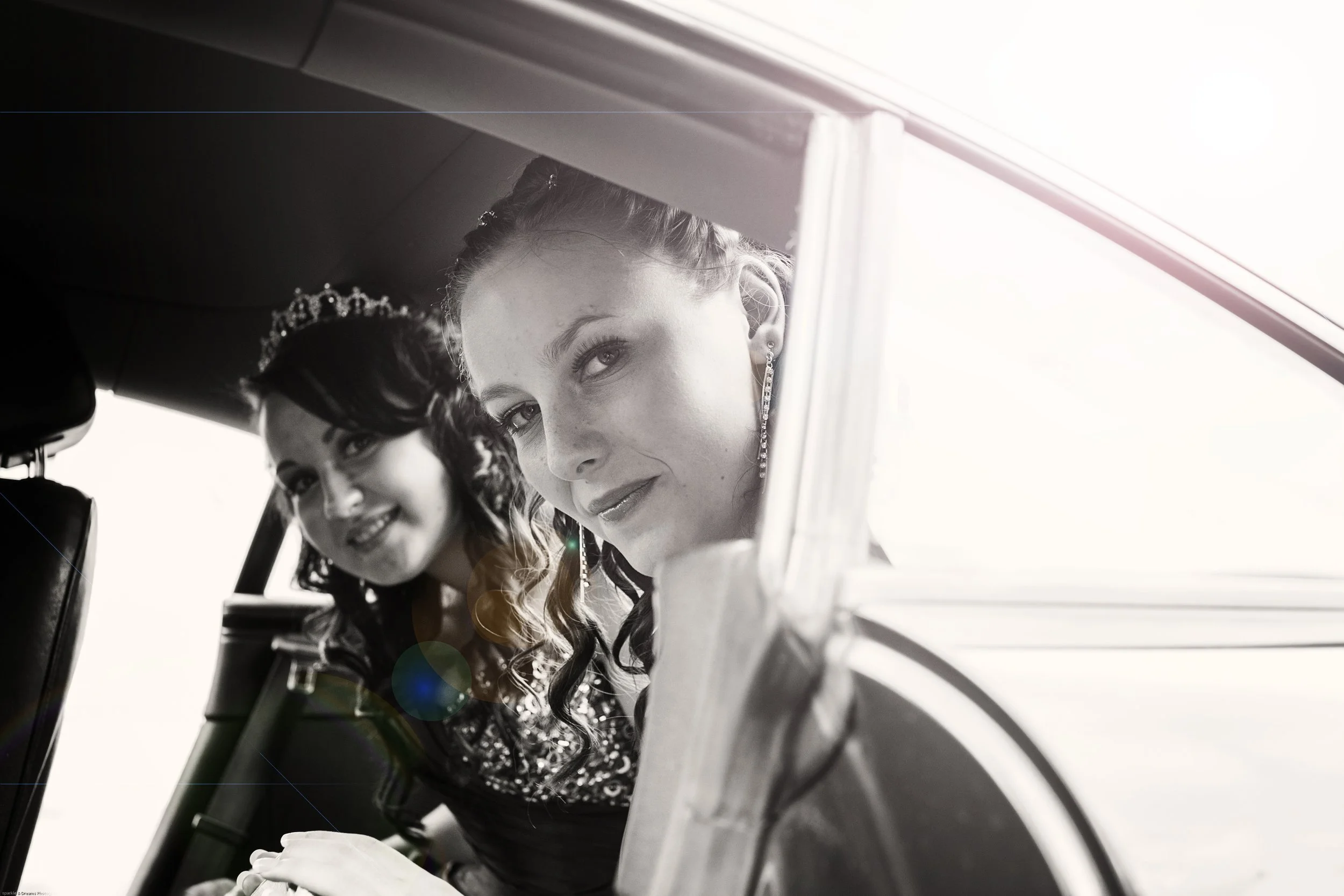 Two women in formal attire leaning out of a car window, smiling at the camera. One woman is wearing a tiara and earrings, and the other is wearing a crown or tiara with wavy hair.