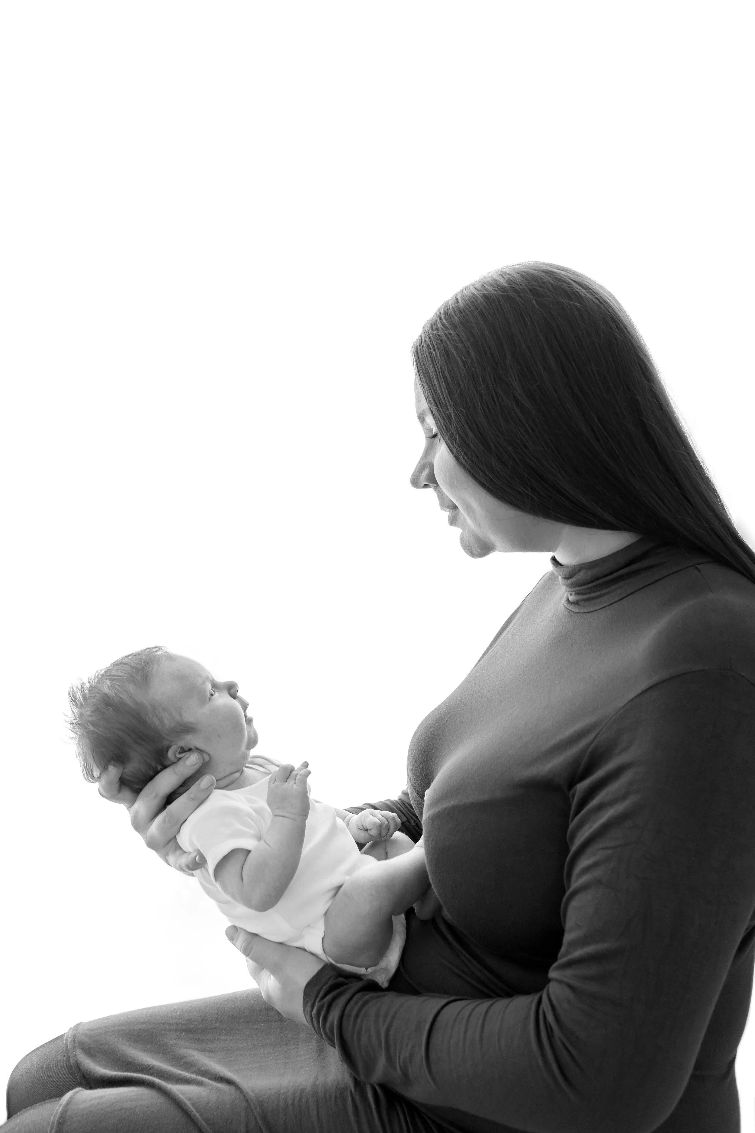 A woman with long dark hair holds a newborn baby in her arms, both looking at each other, against a plain white background.