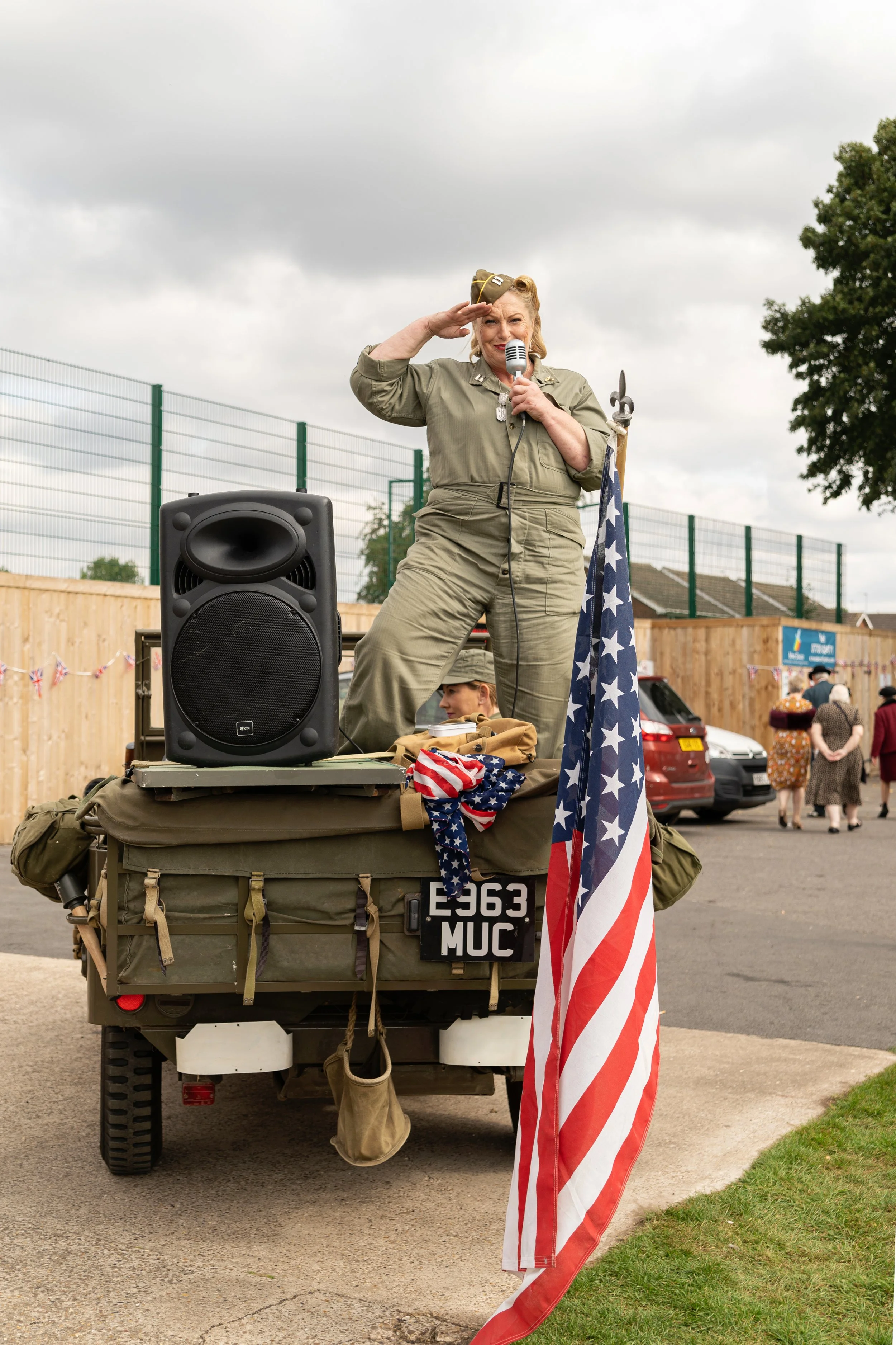 A woman dressed in vintage military uniform standing on a small military vehicle, saluting and speaking into a microphone. The vehicle is decorated with American flags, and there are people in the background attending an outdoor event on a cloudy day.