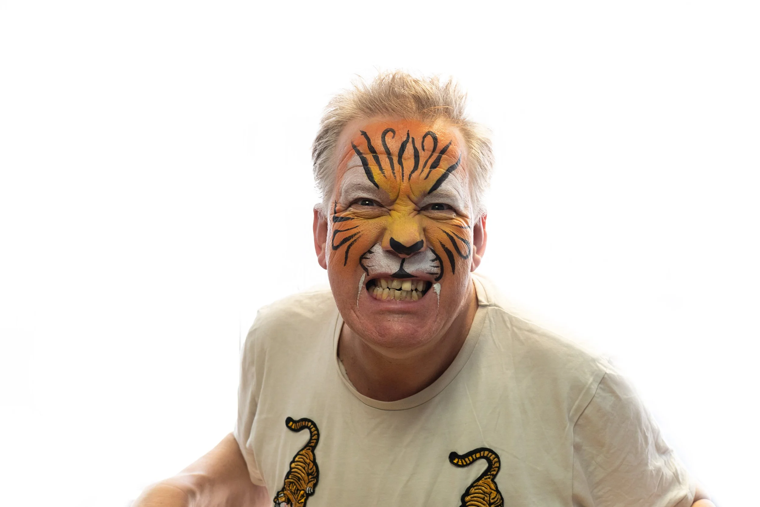 Man with tiger face paint making a fierce expression, wearing a light-colored T-shirt with tiger related designs, against a plain white background.