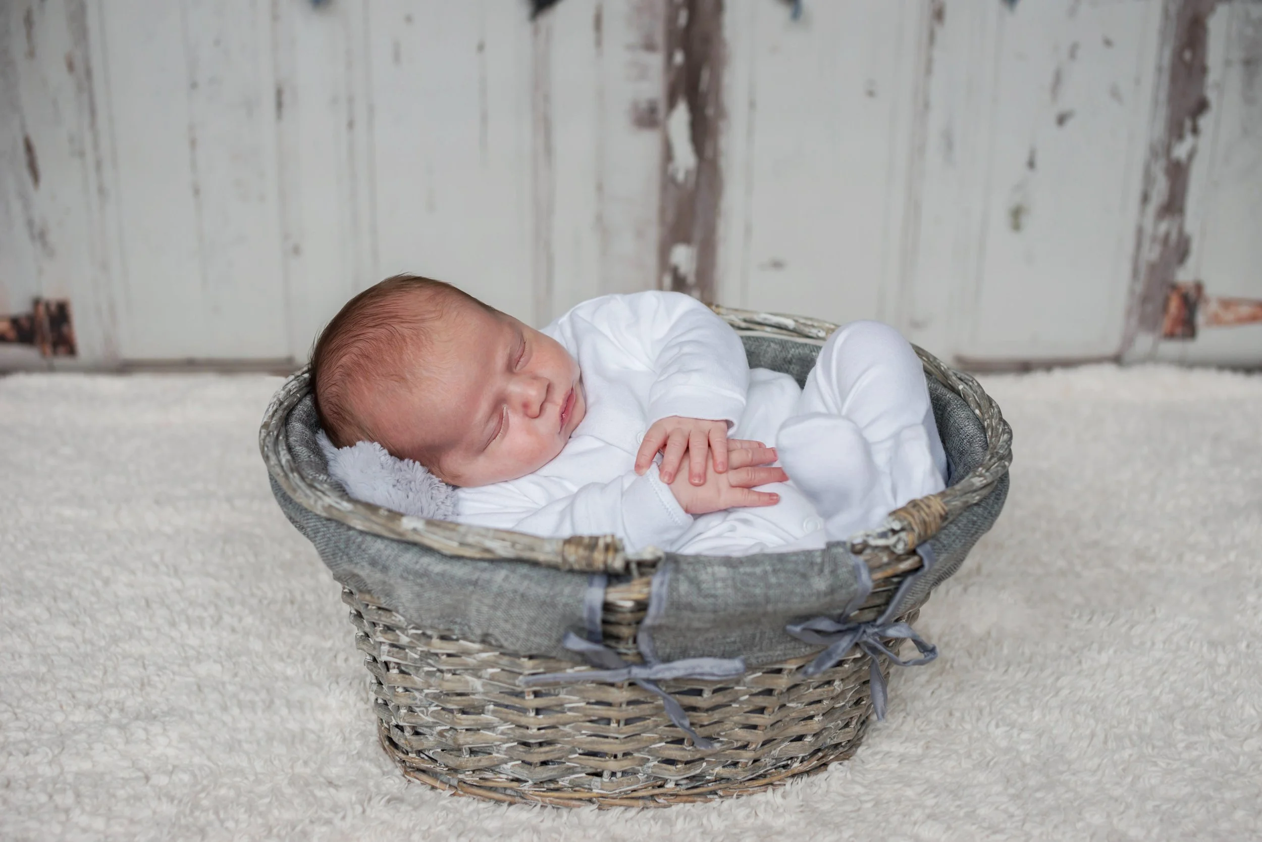 A sleeping baby in a gray wicker basket with a white blanket, on a cream-colored carpet, with a rustic wooden background.