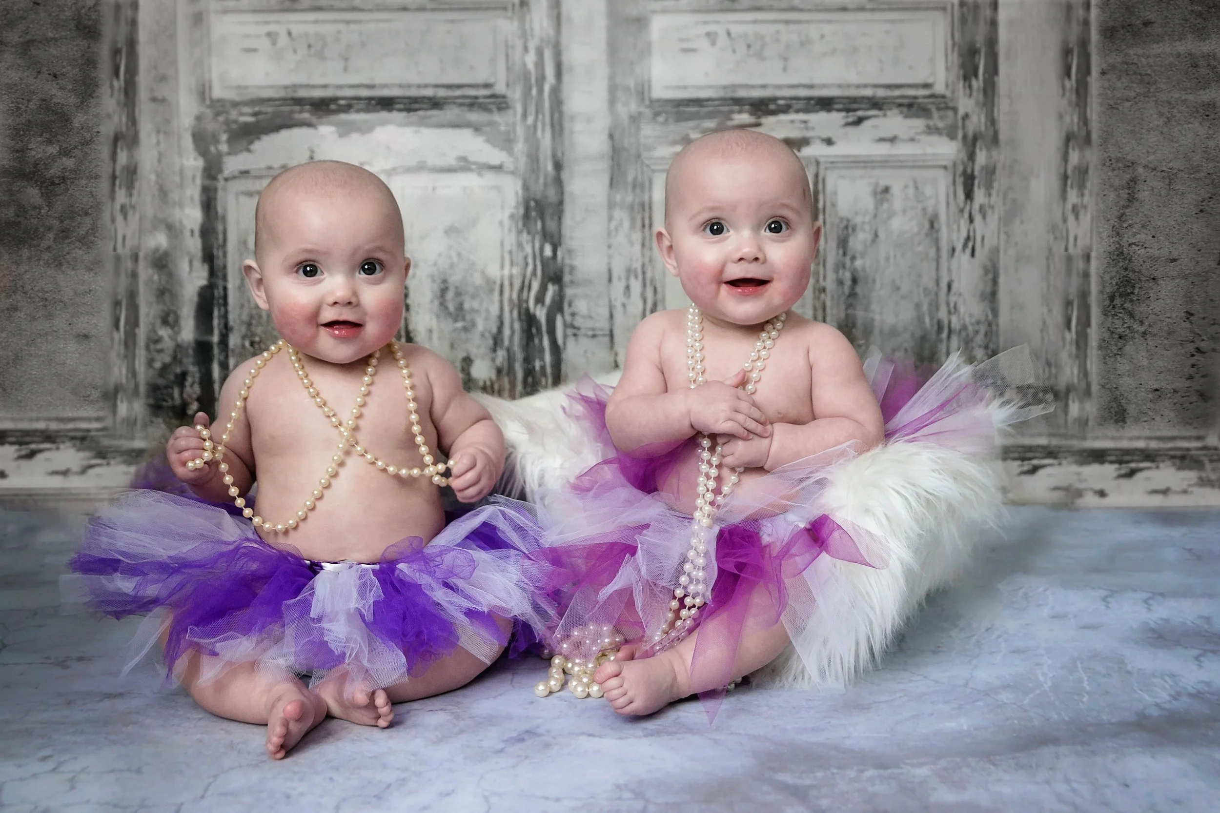 Two babies sitting on a soft surface, dressed in purple Tutu skirts with pearl necklaces, in front of a rustic wooden panel backdrop.