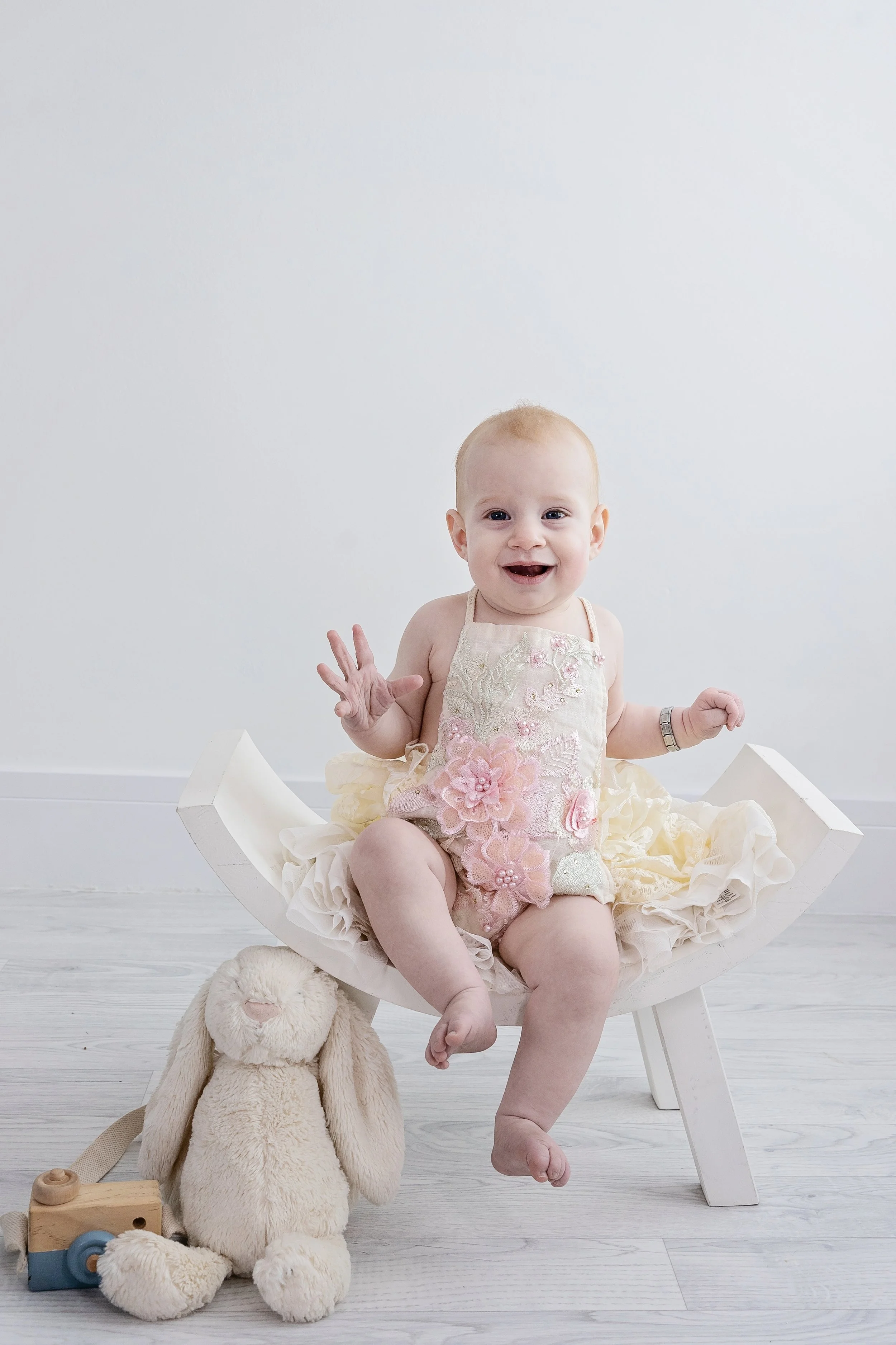 A smiling baby girl with blonde hair sitting on a small white wooden bench, wearing a pastel pink floral dress, with a plush bunny toy and a wooden camera toy on the floor beside her, set against a white wall.