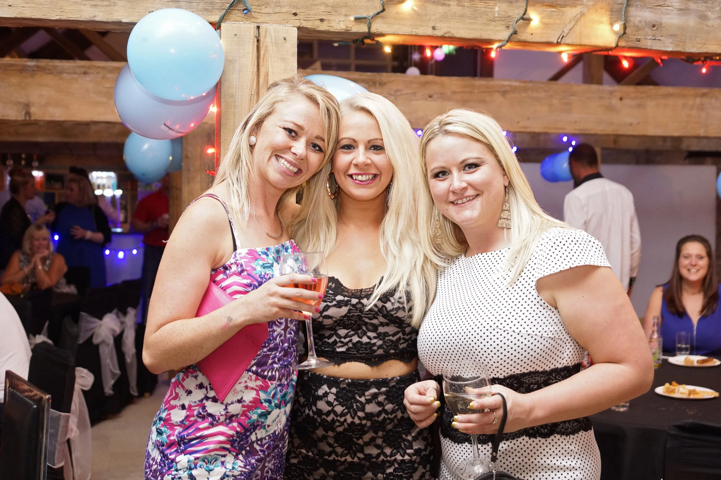 Three women smiling and holding drinks at a celebration or party, with balloons and string lights in the background.
