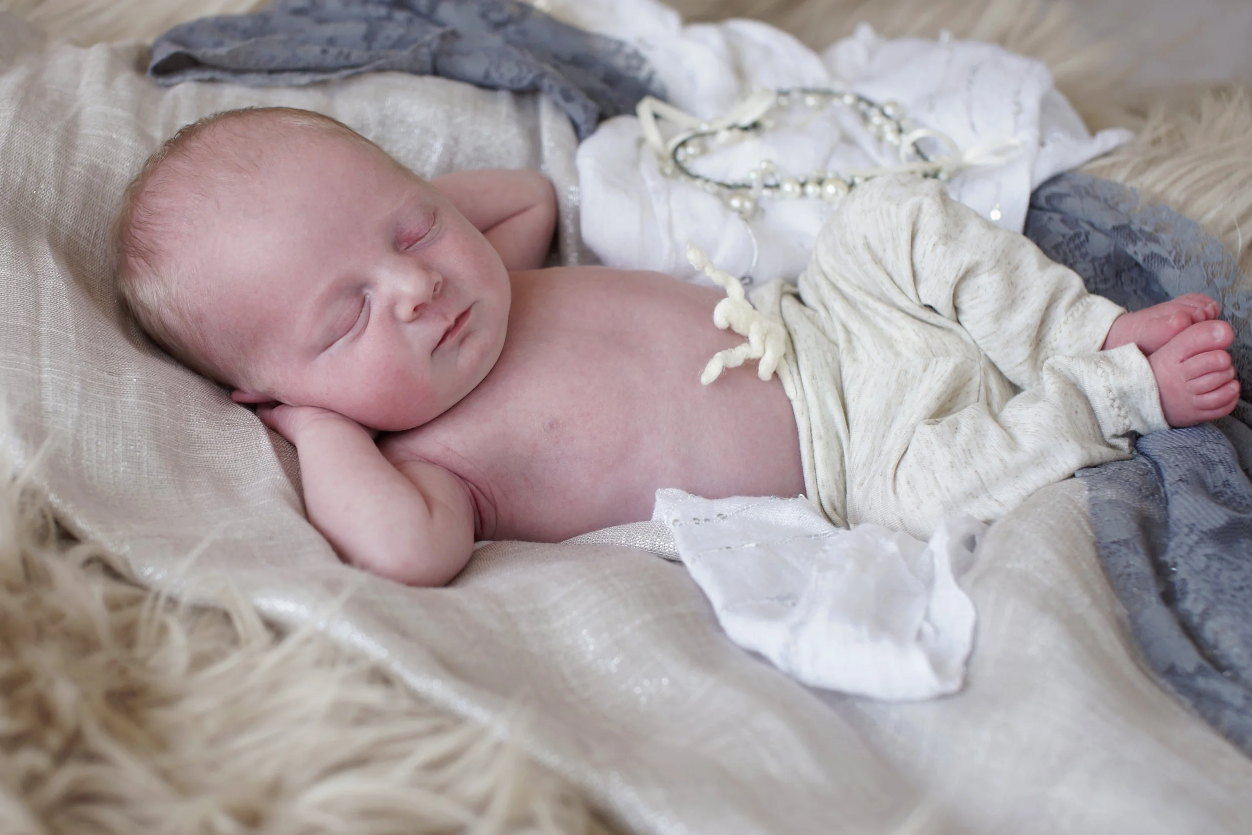 A newborn baby with light skin sleeping on a soft, light-colored blanket surrounded by jewelry and fabric.
