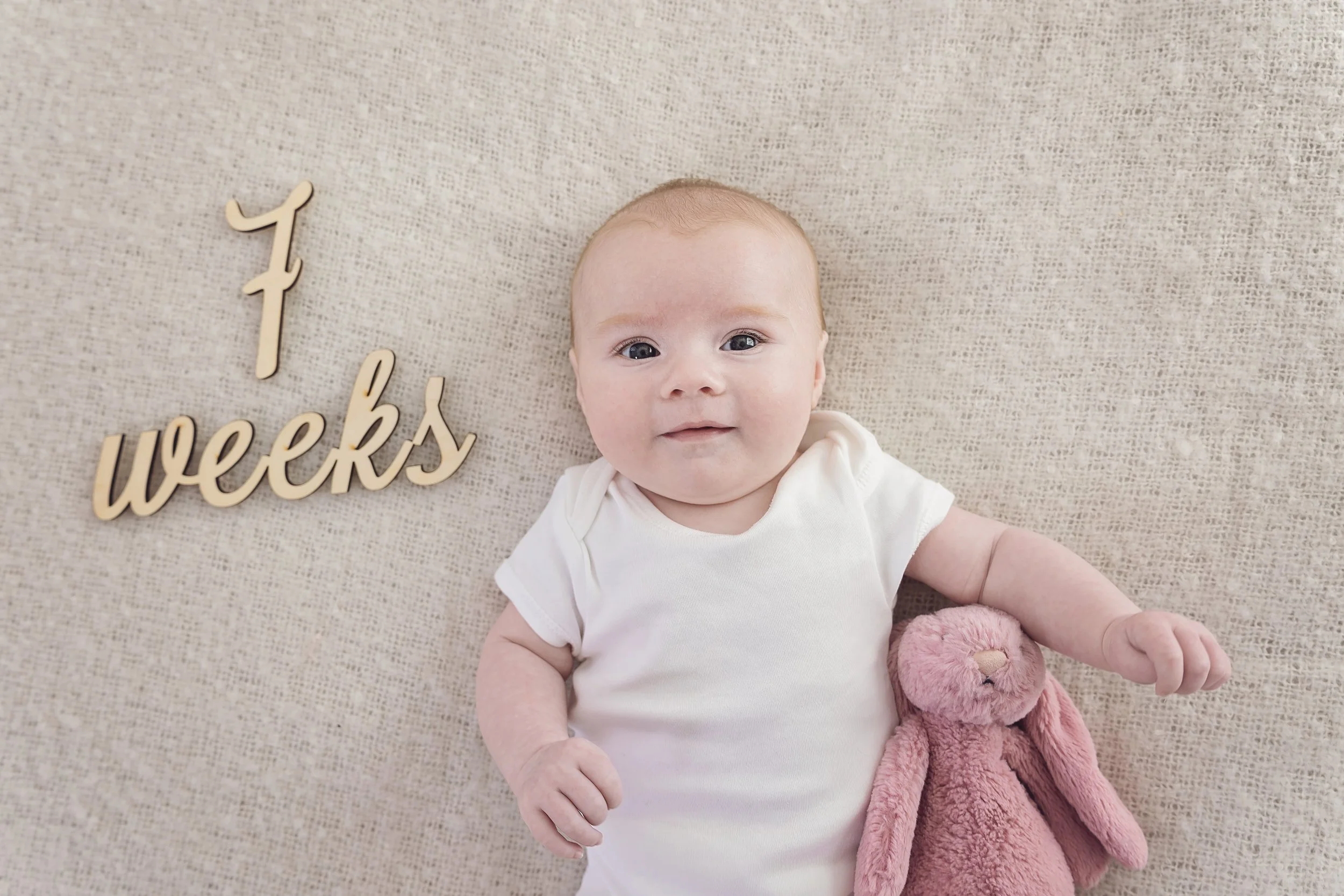 A smiling baby with blue eyes and light hair lying on a beige textured surface, holding a pink stuffed bunny, next to a wooden sign that reads 'f weeks' indicating a baby's age.