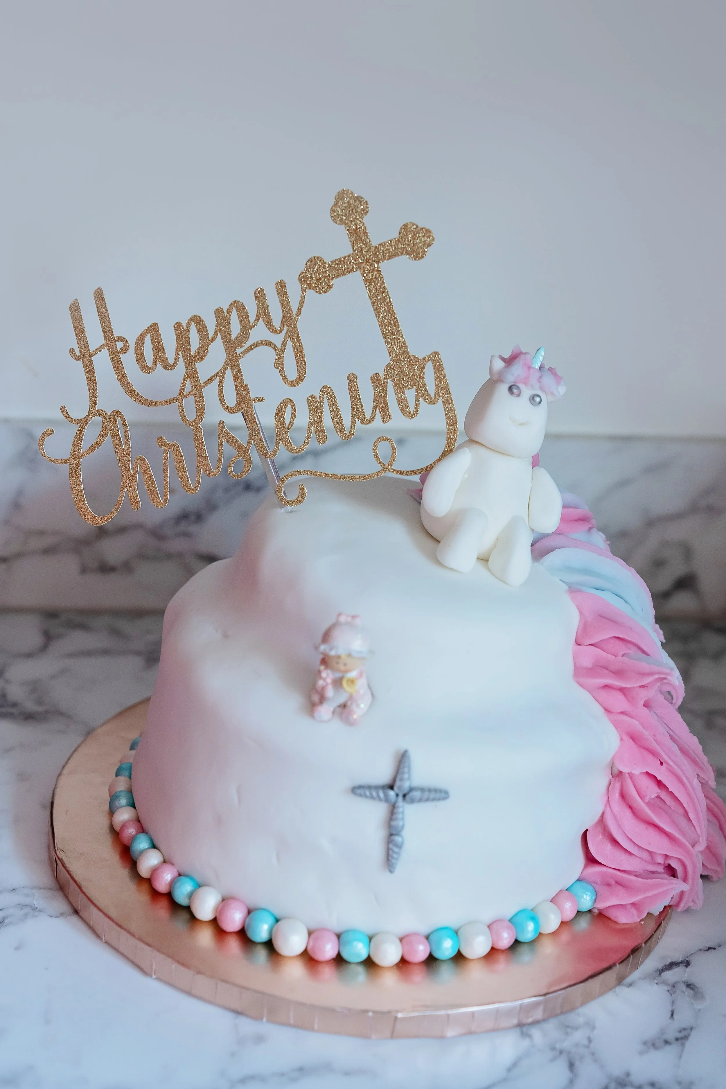 A decorated birthday cake with white fondant, pink and blue frosting accents, a unicorn figurine, and a glittery gold topper that reads 'Happy Christening' with a cross, placed on a marble surface.