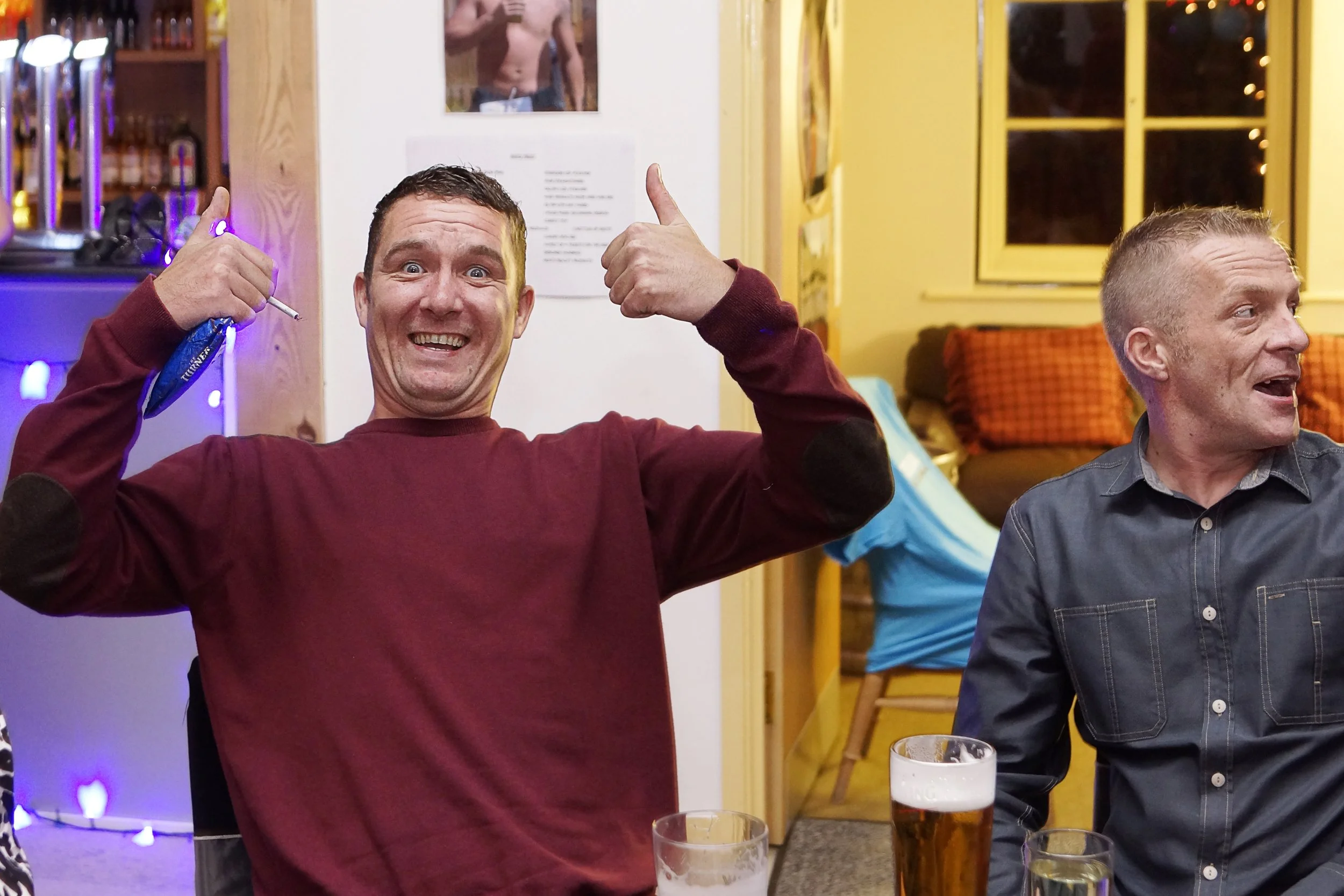 A man in a maroon sweatshirt is smiling and giving a thumbs-up, holding a vape device, while sitting at a table with drinks; another man in a gray shirt is sitting beside him, looking to the side, in a warmly lit indoor setting.