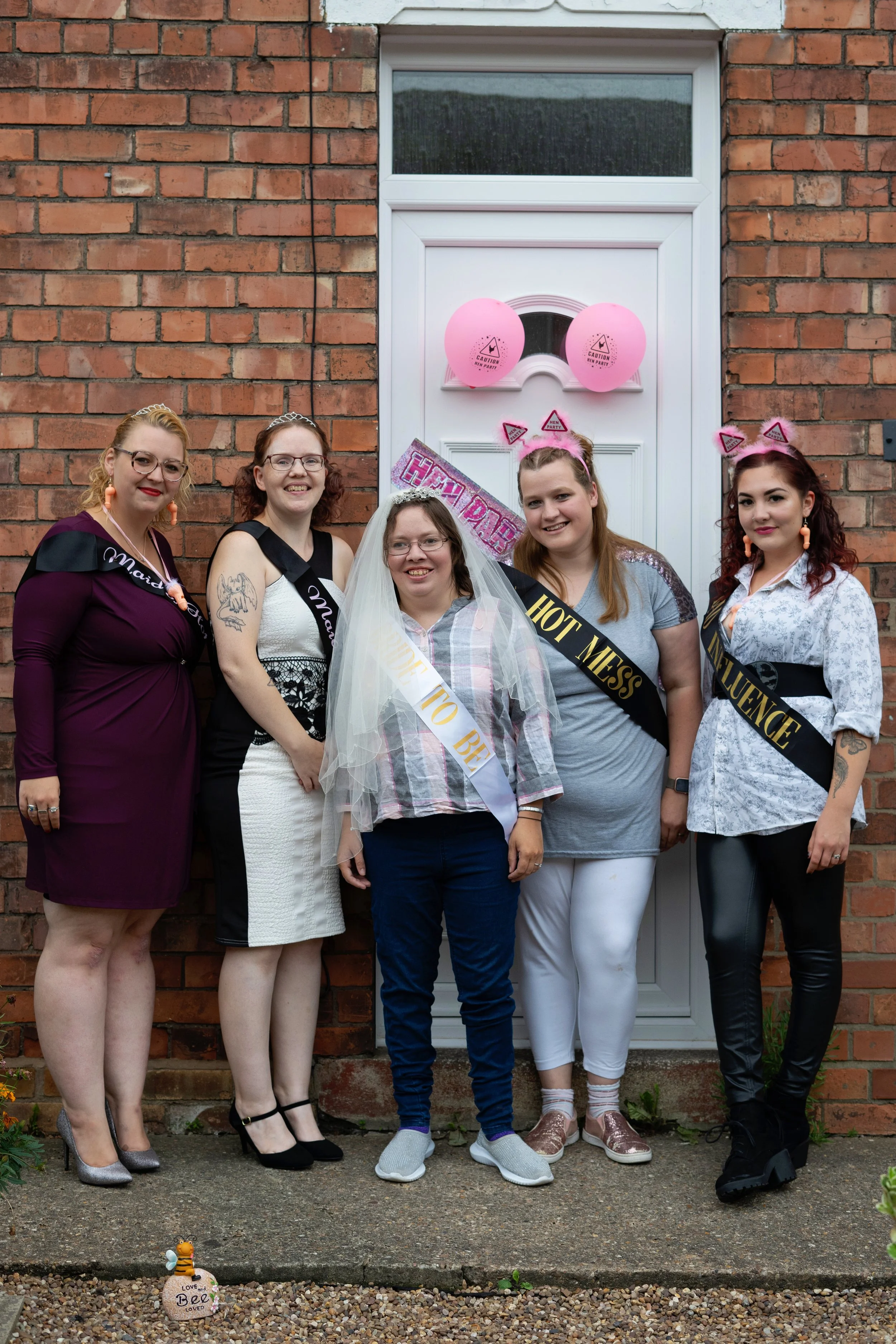 Group of five women celebrating bachelorette party with sashes, tiaras, and pink decorations outside brick house.