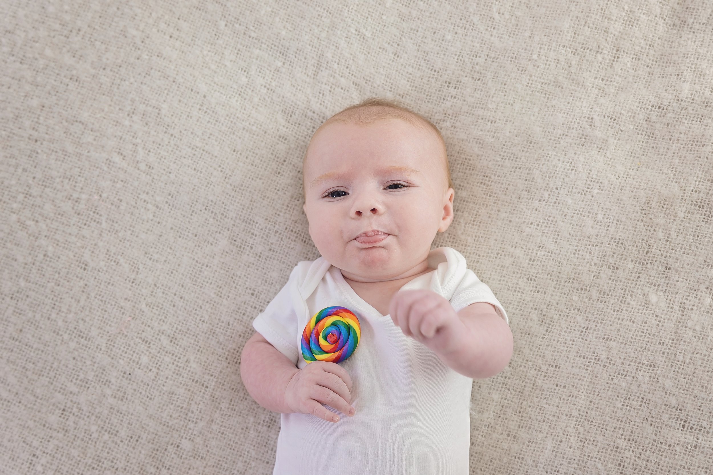 Baby lying on beige blanket holding a colorful rainbow lollipop, making a playful face with tongue slightly out.