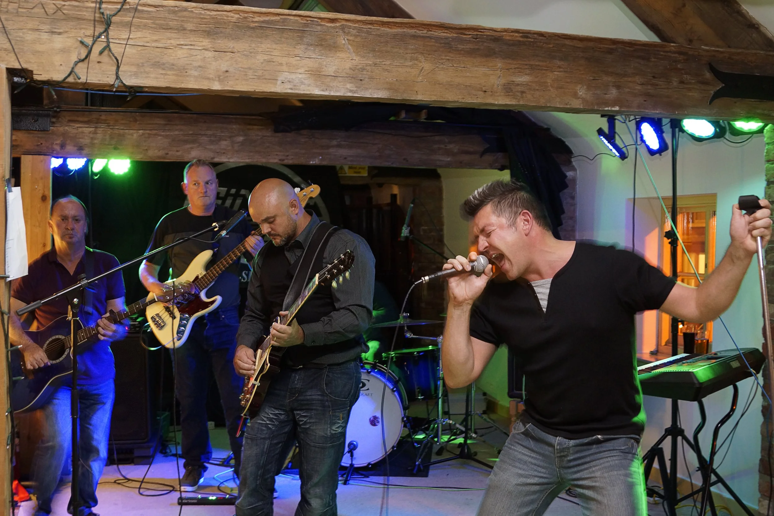 A band of four men performing live music on stage with guitars, drums, keyboard, and singing. Colorful stage lights illuminate the scene.