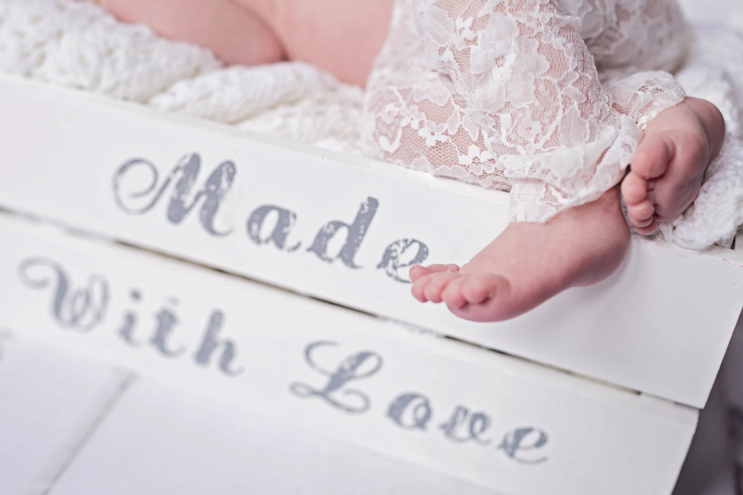 Close-up of a newborn baby lying on a white wooden box with the words 'Mad' and 'Witch' painted on it. The baby is wearing a white lace outfit, with only the legs and feet visible.