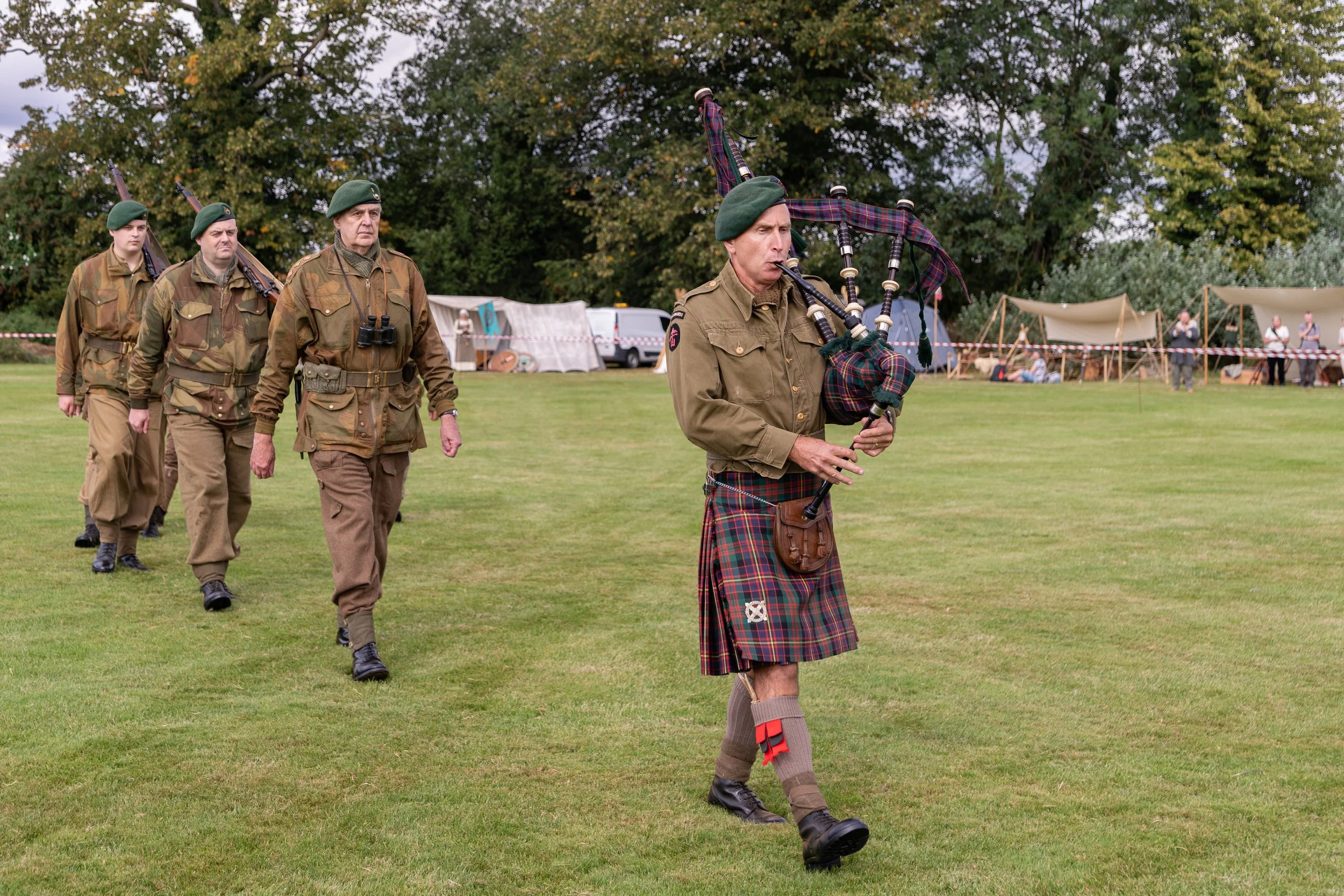 A group of four men dressed in mid-20th century military uniforms, with the front man playing the bagpipes, walking across a grassy field during a historical reenactment event.