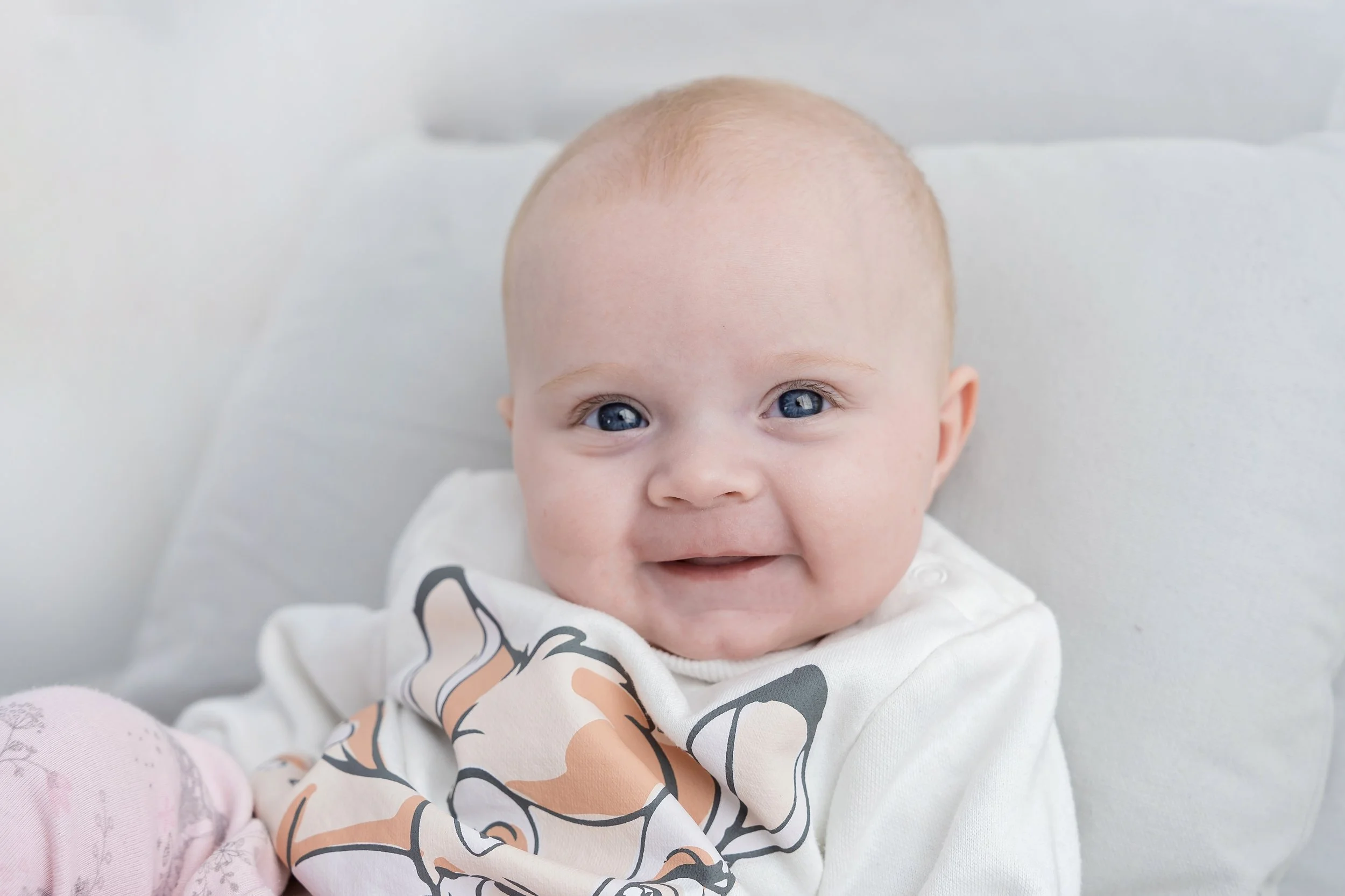 A smiling baby with blue eyes lying on a white pillow, wearing a white shirt with a cartoon dog print and pink pants.