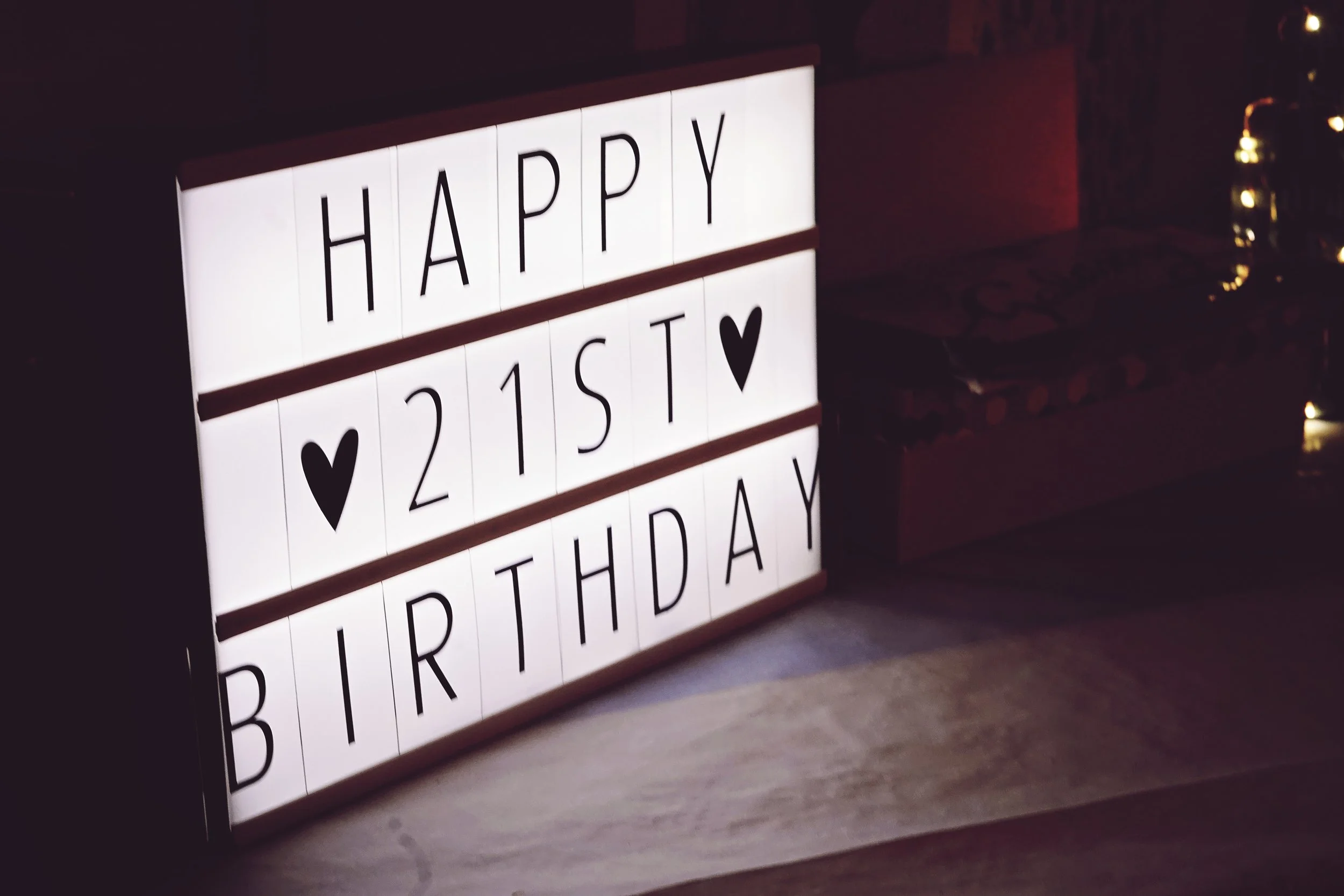 Lightbox sign with black letters on white background reading "HAPPY 21ST BIRTHDAY" with two black heart symbols, placed on a table in a dimly lit room.