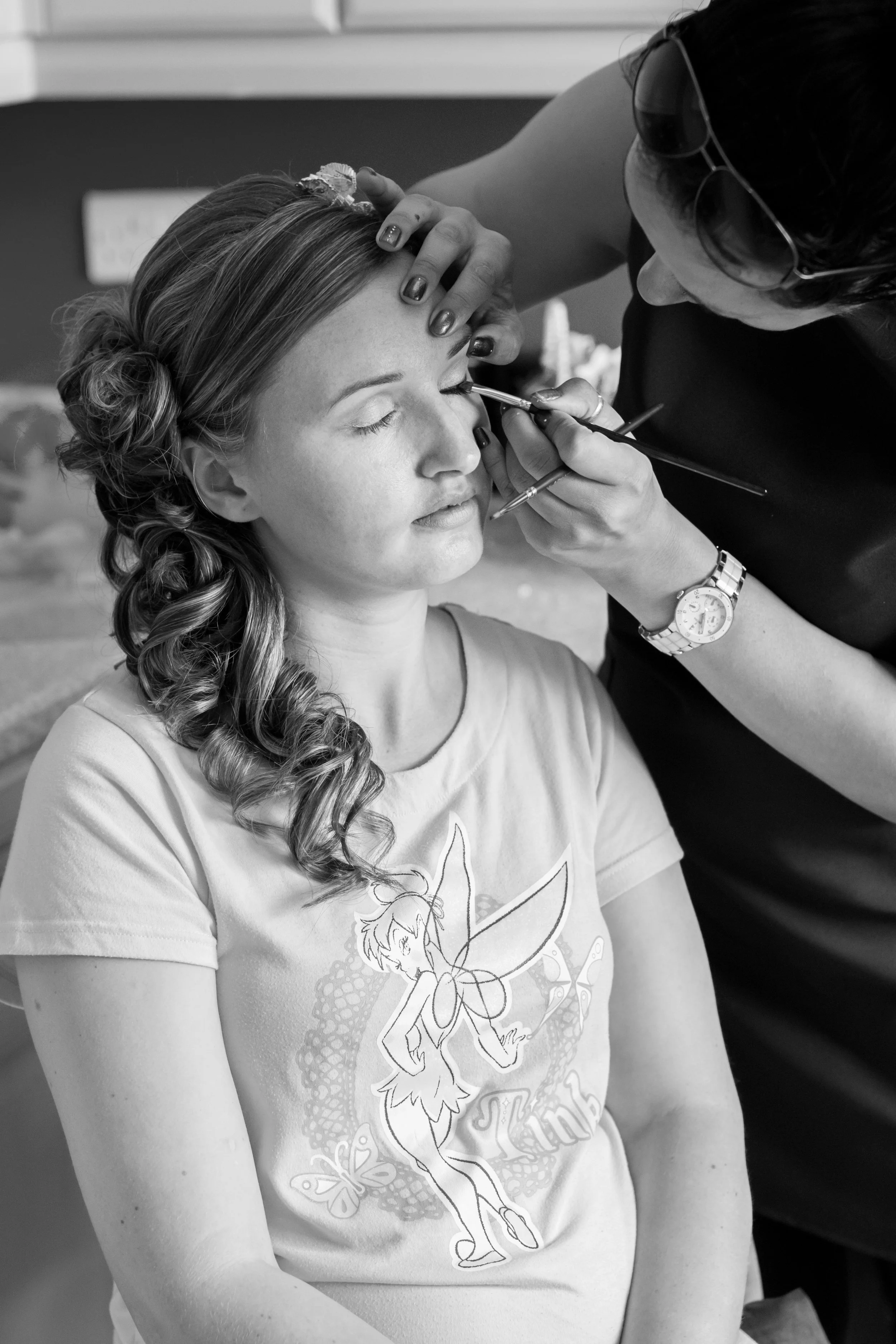 A woman getting her makeup done by a makeup artist, who is applying makeup around her eyes in a backstage setting.