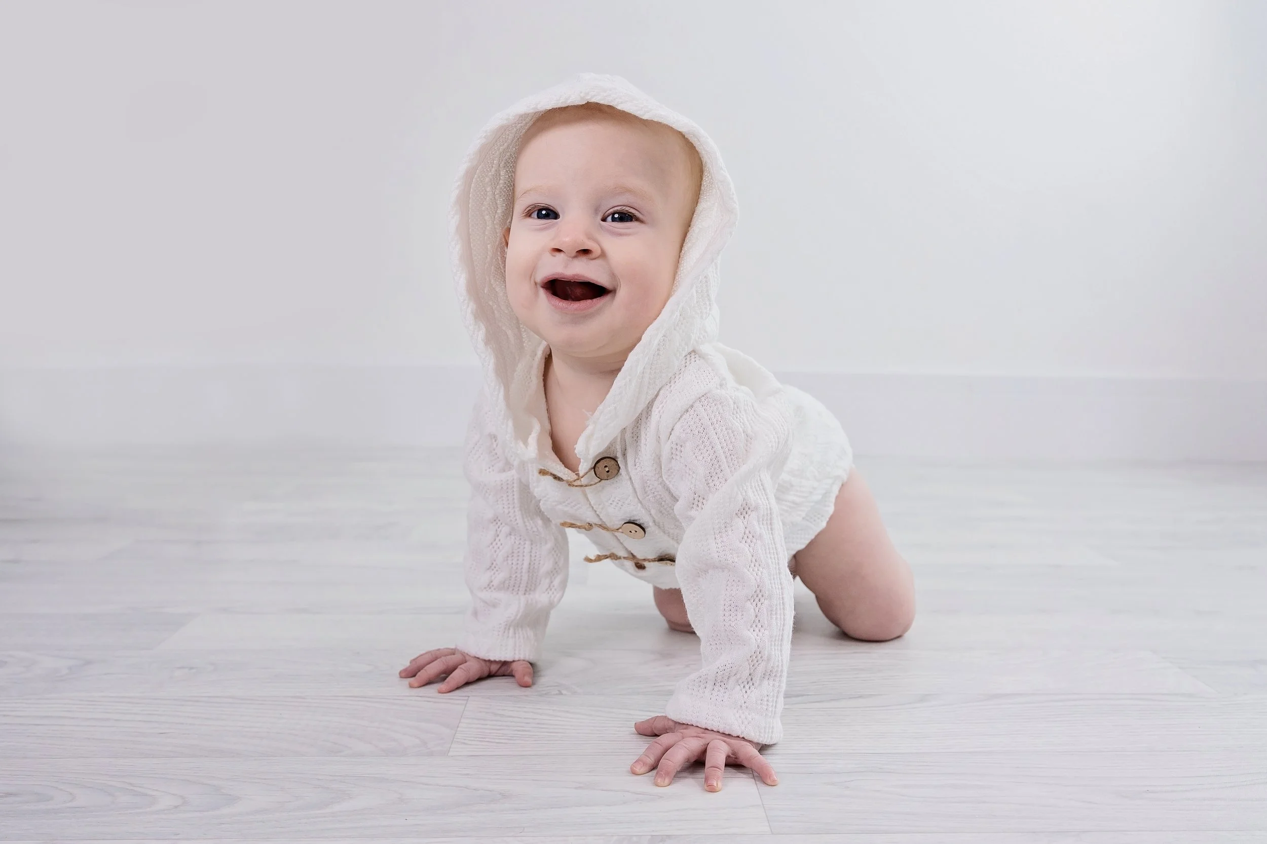 A baby crawling on a light wood floor, smiling and wearing a white knitted hoodie and matching cardigan with wooden buttons.