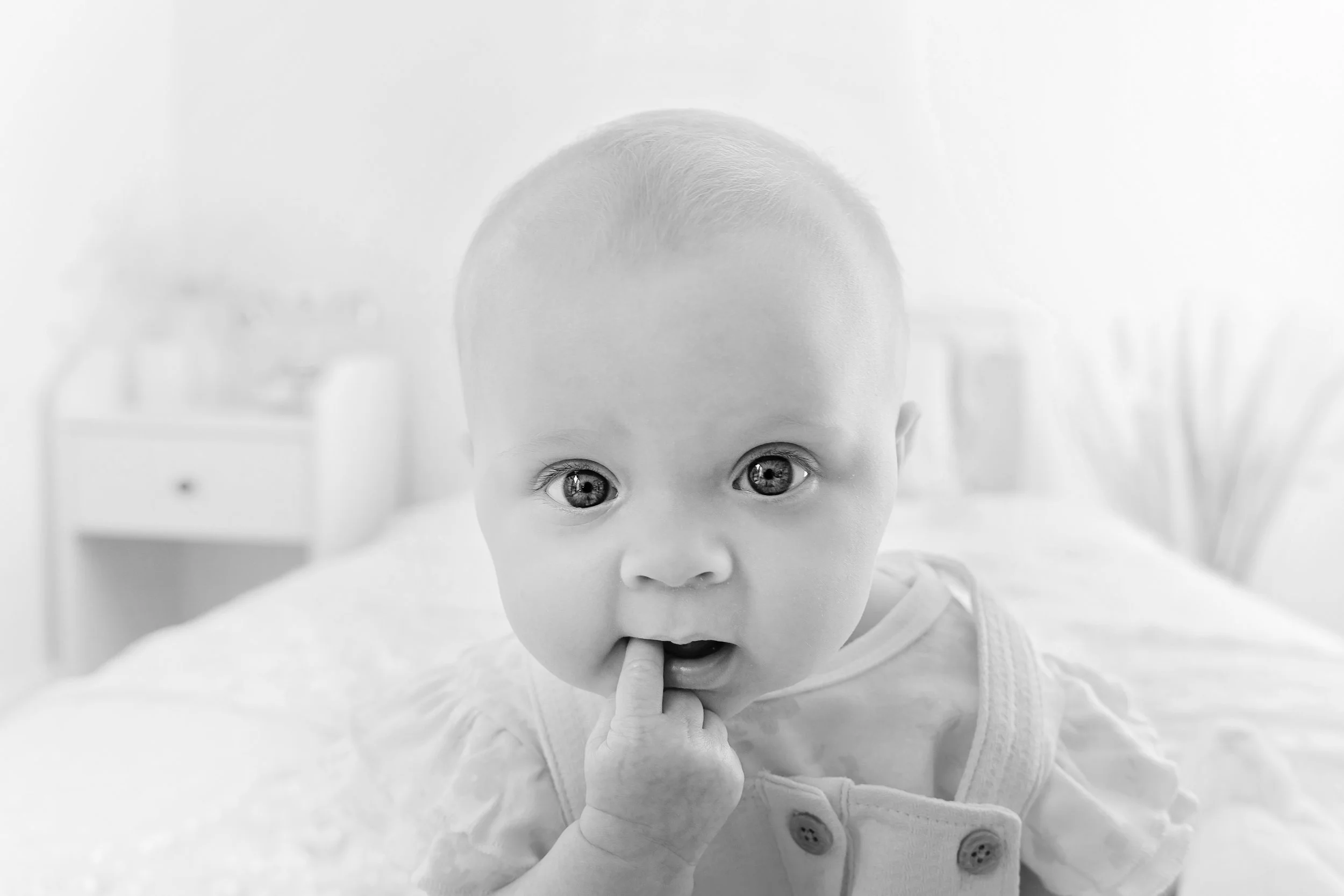 Black and white photo of a baby with big eyes, lying on a bed, with one finger in their mouth, looking directly at the camera.