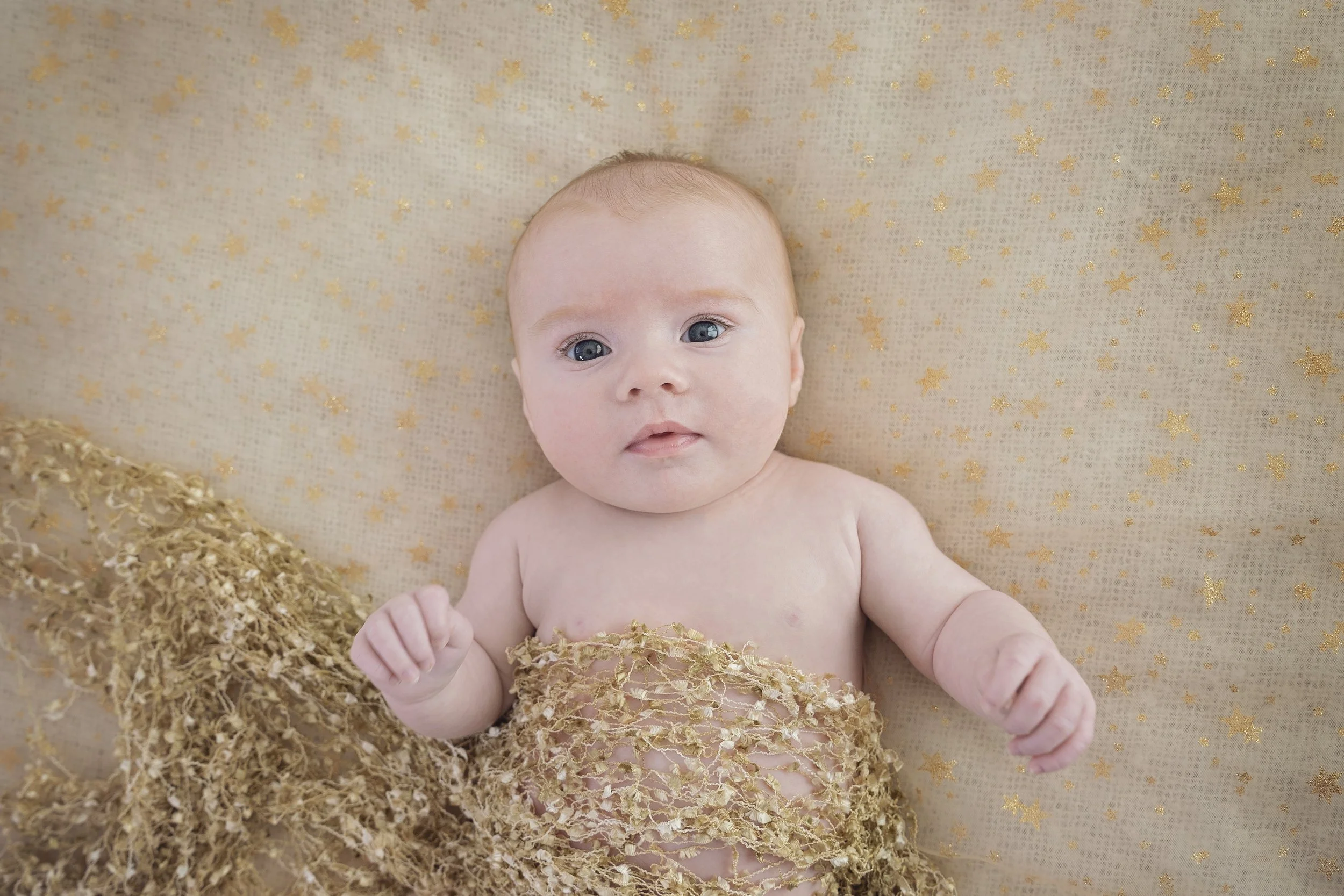 A baby with blue eyes lying on a beige blanket with gold stars, partially covered with a beige lace fabric.