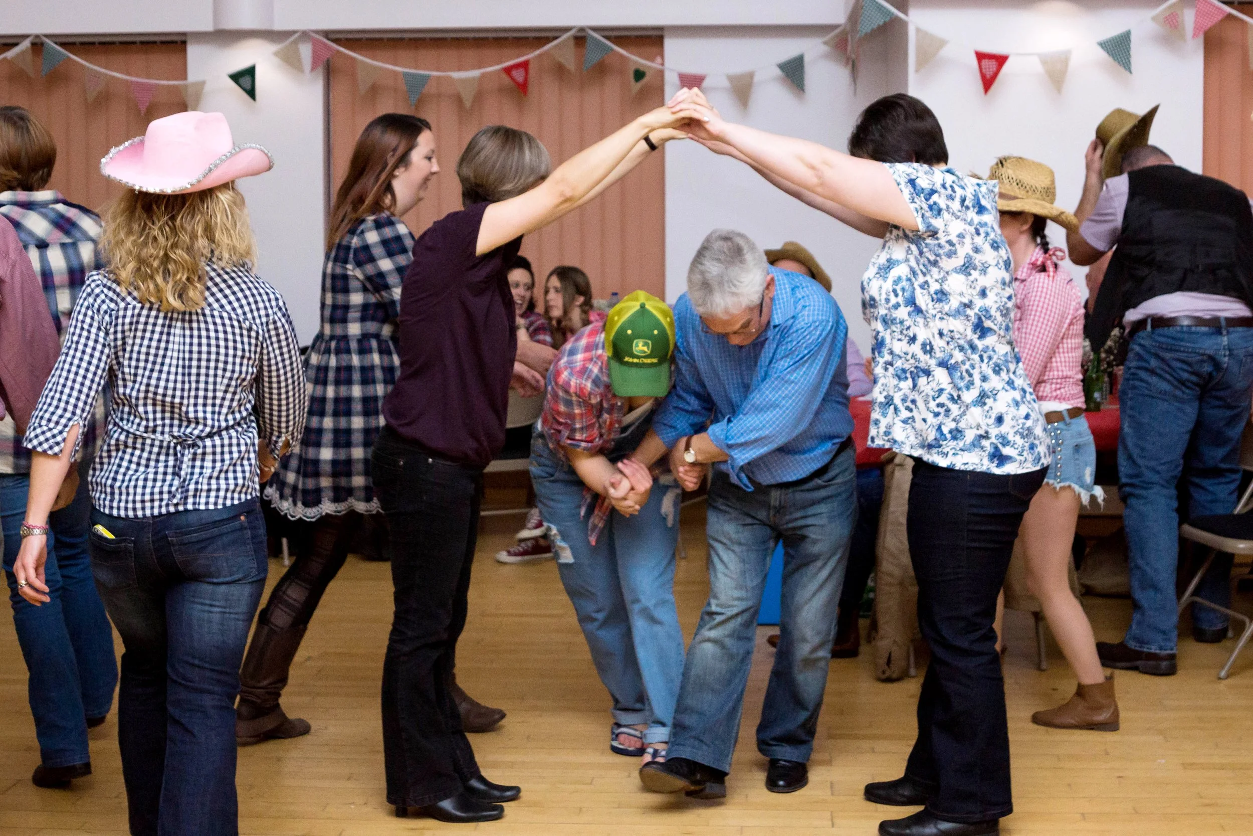 People dancing in a line at a country-themed party, with some wearing cowboy hats and plaid shirts, and decorations like bunting hanging from the ceiling.