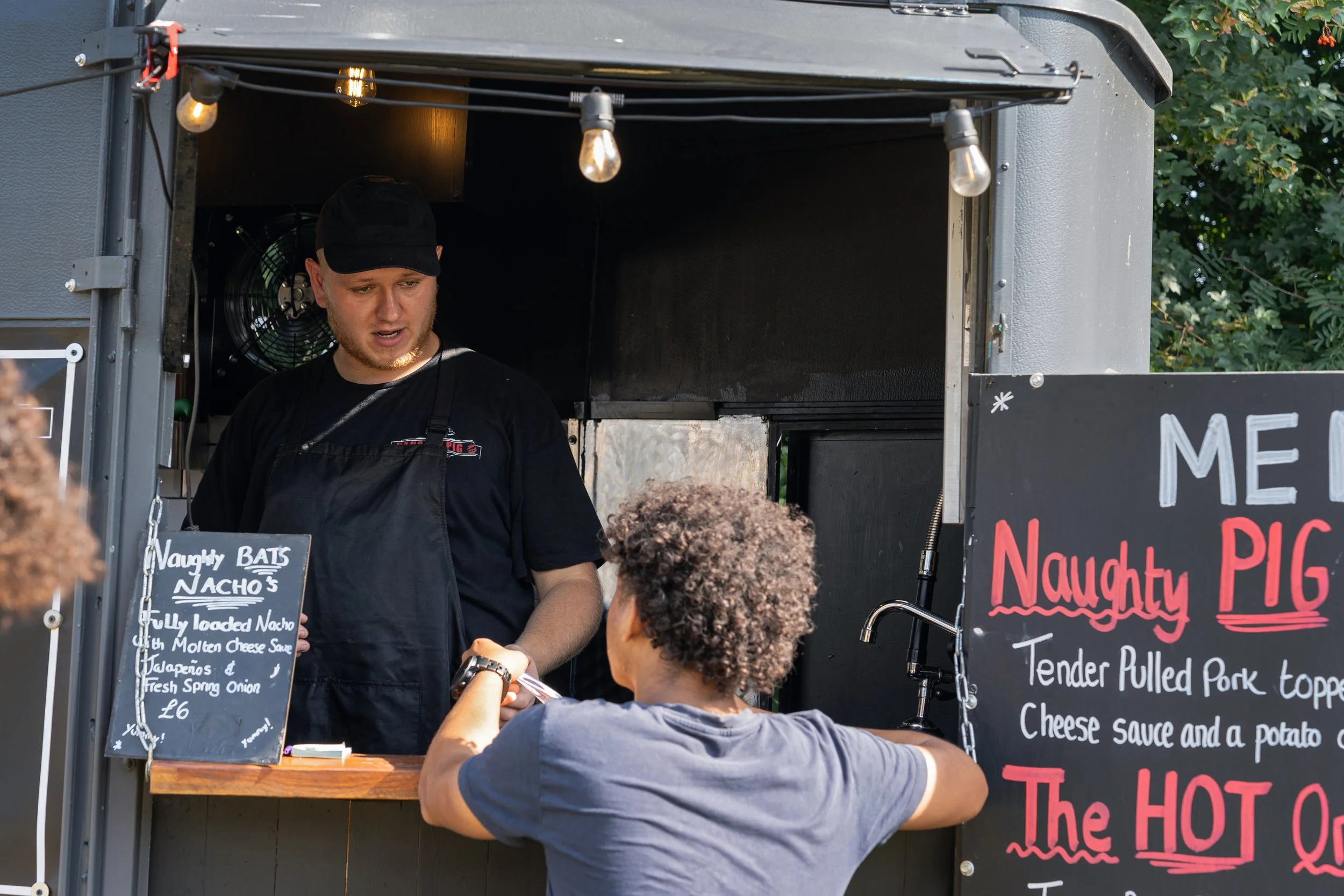 A food stand with a man selling pork topped nachos to a customer with curly hair. The stand displays chalkboard menus with food items and prices, and has string lights hanging from the top.