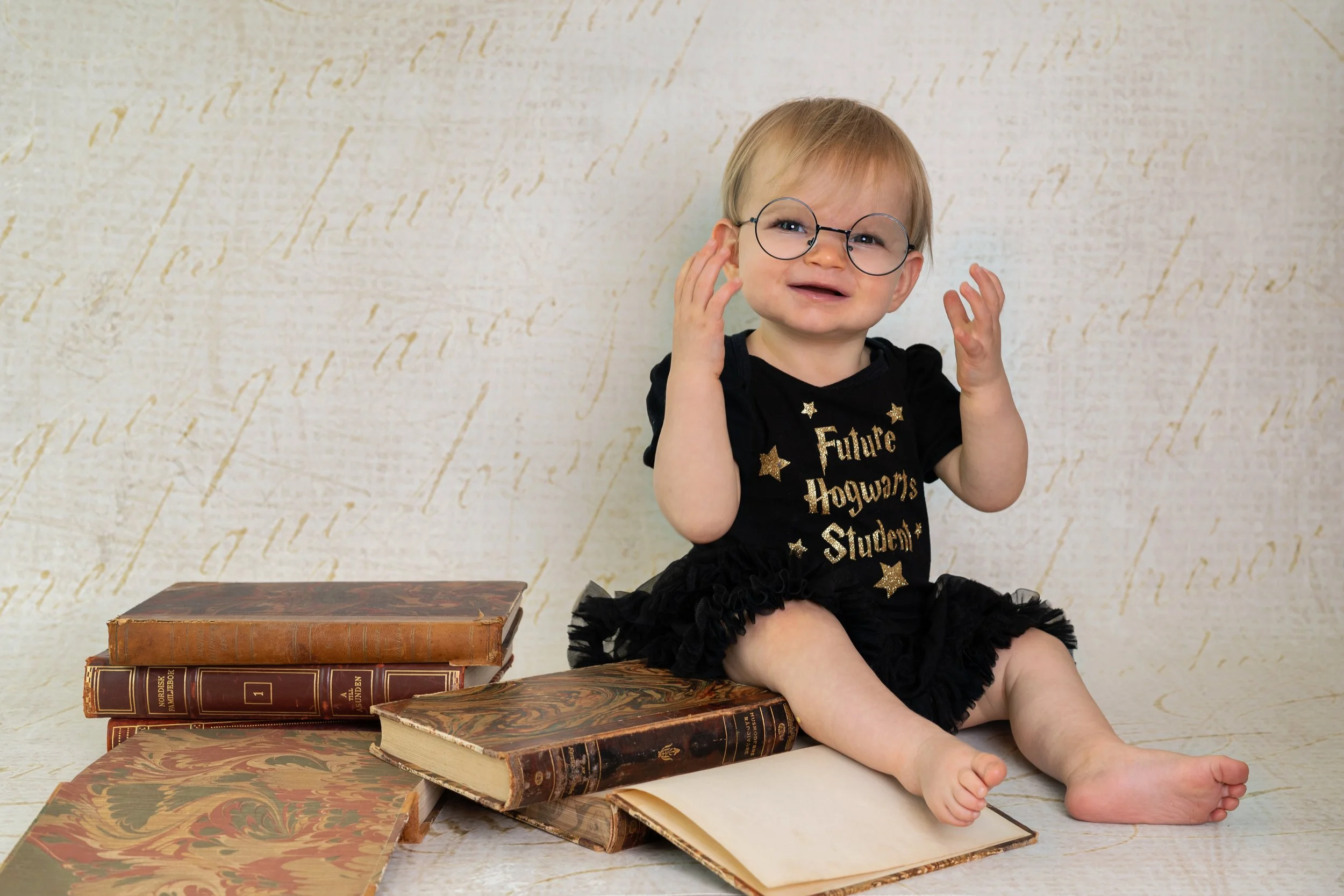 A toddler wearing glasses and a black dress with gold glitter text that reads 'Future Hogwarts Student' is sitting on the floor with old books around her, holding her head with a distressed expression.