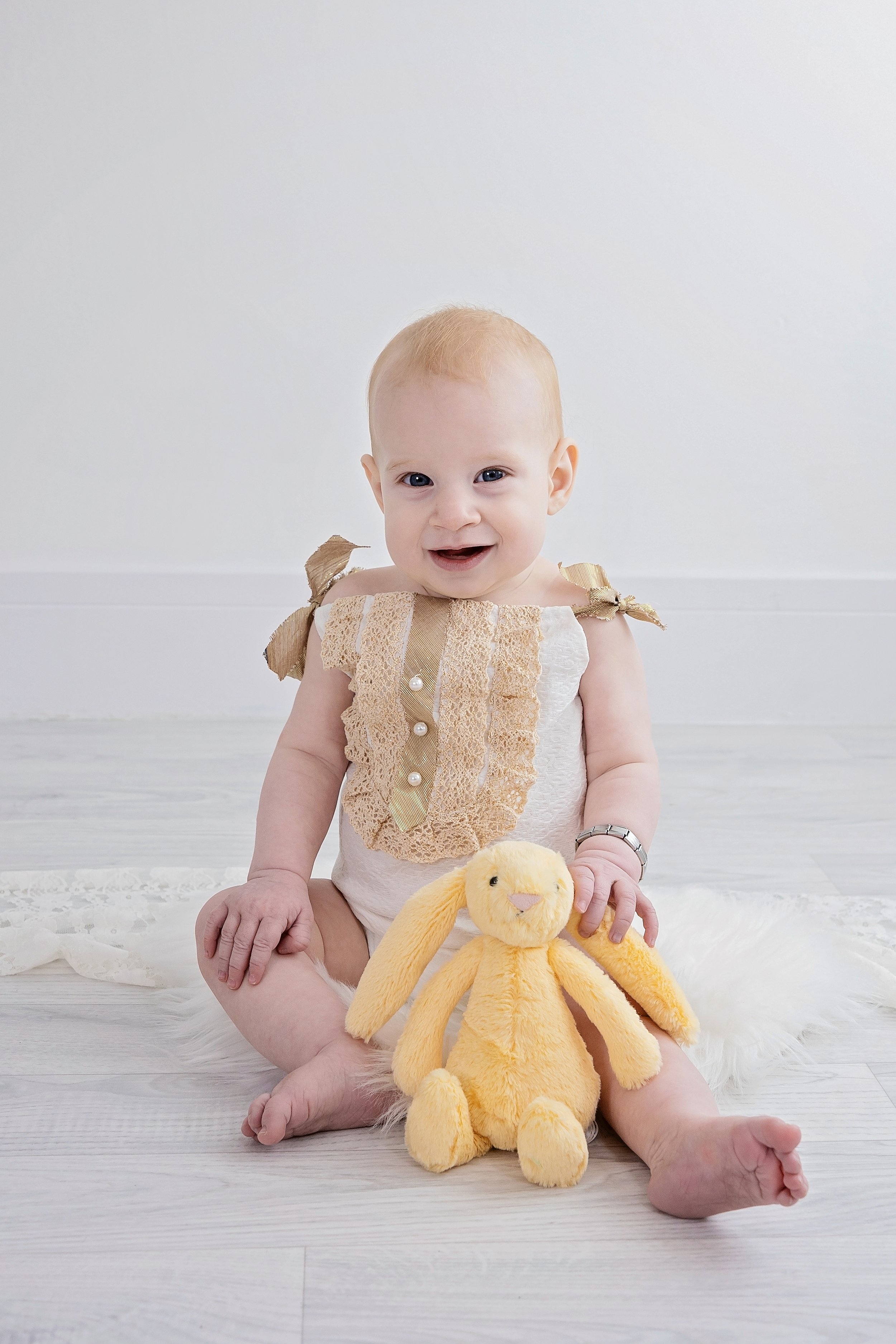A smiling baby girl with blonde hair, sitting on a white floor, holding a yellow stuffed bunny.