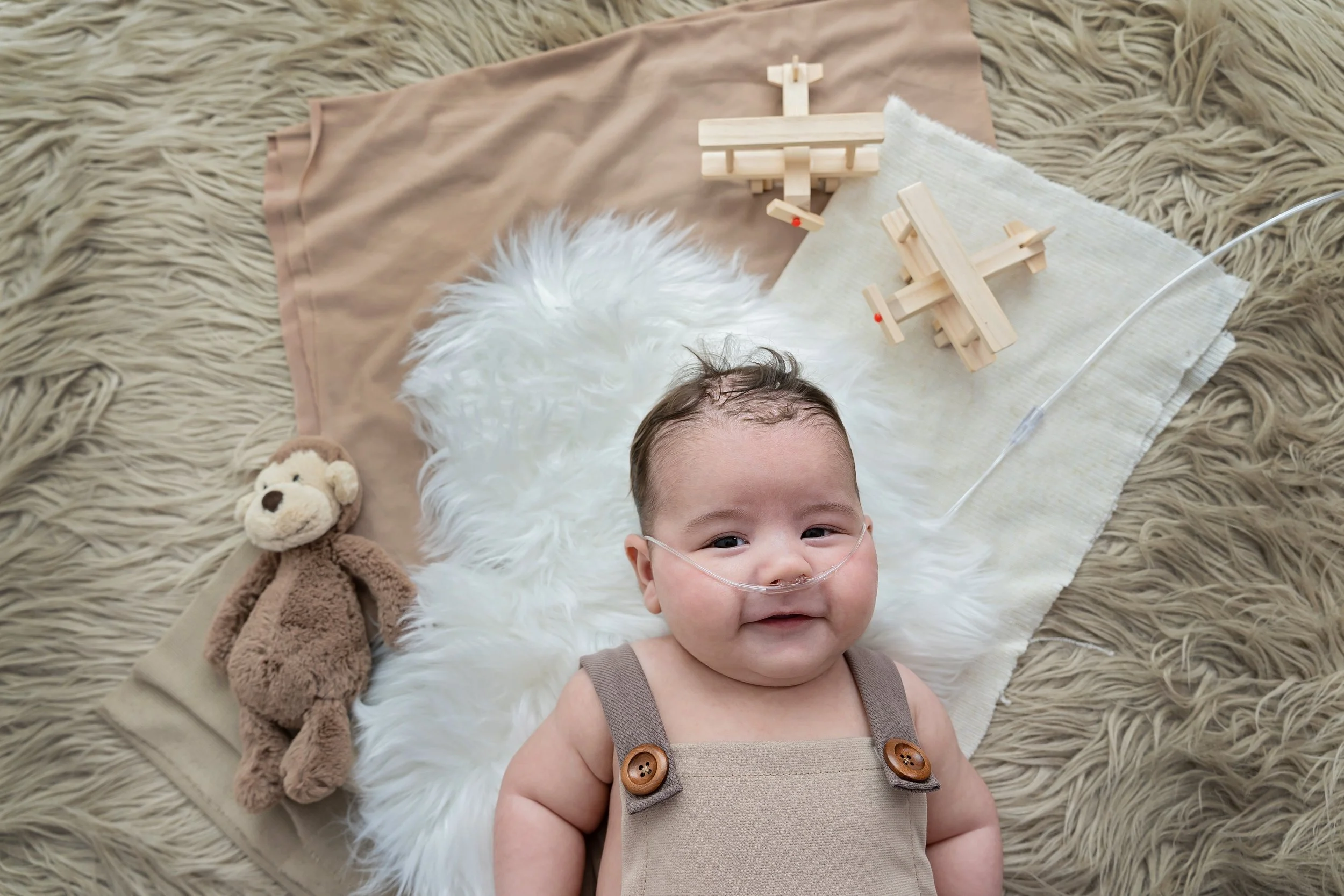 Smiling baby with a nasal oxygen tube lying on a soft blanket with teddy bear, wooden toy helicopters, and a beige pillow surrounding him.