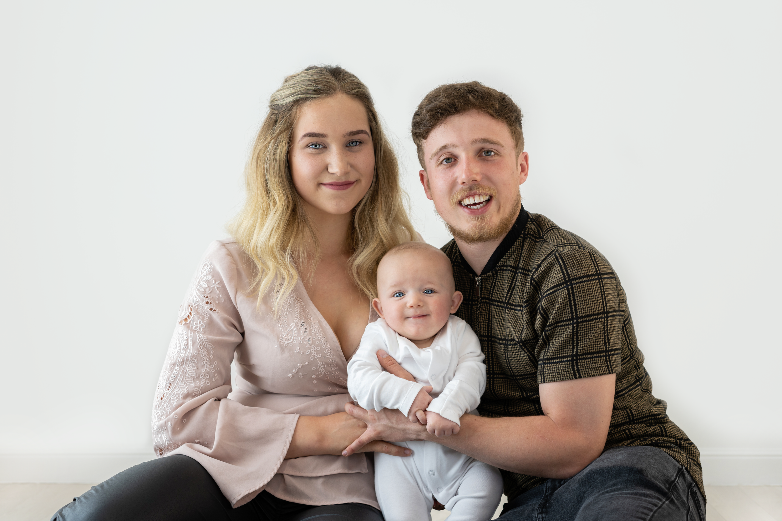 A young family of three sitting against a white wall, with the mother on the left, father on the right, and a baby in the middle, all smiling at the camera.