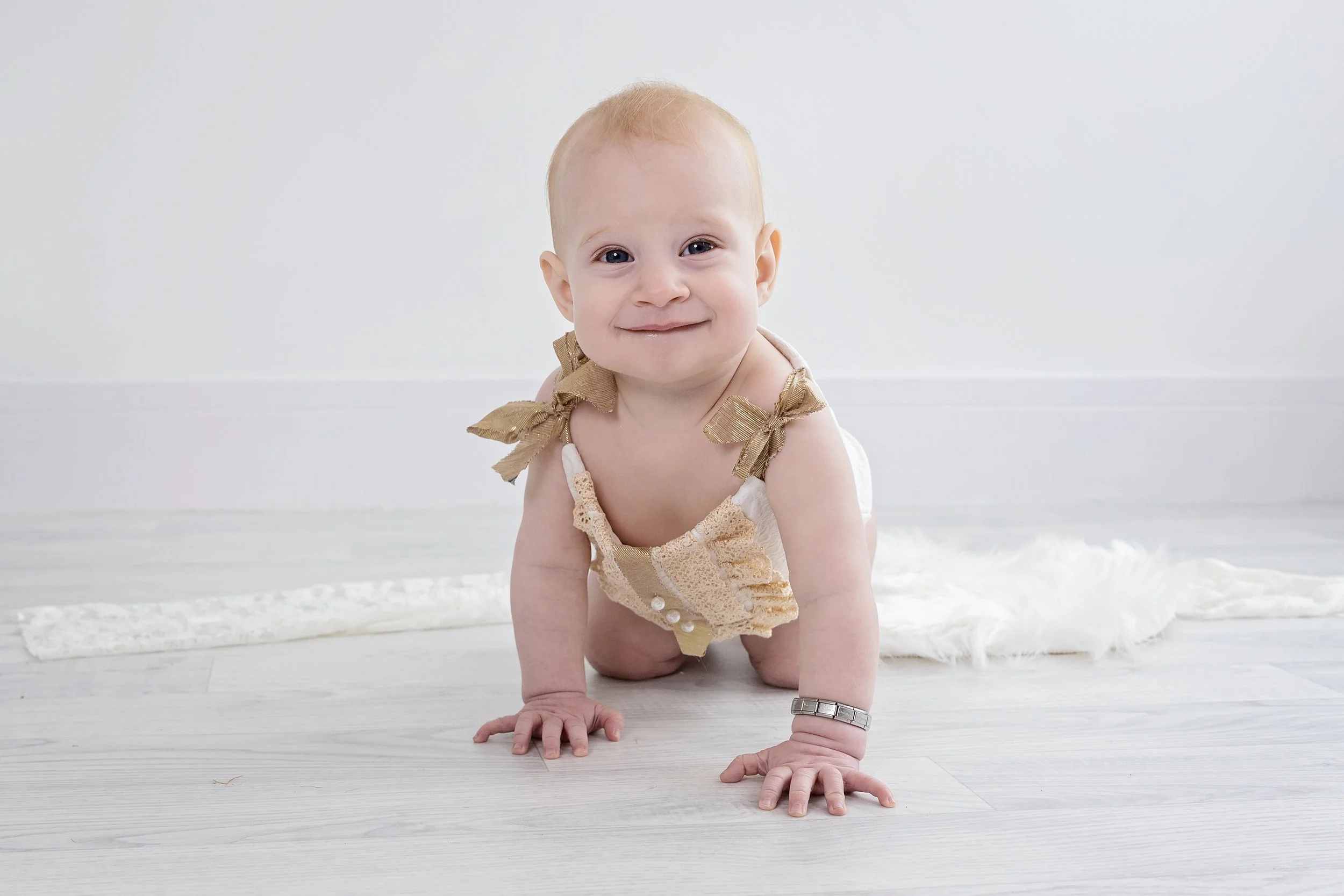 Baby crawling on white floor, smiling, wearing a cream-colored dress with golden bows on the shoulders, with a silver bracelet on the left wrist, and a fluffy rug in the background.