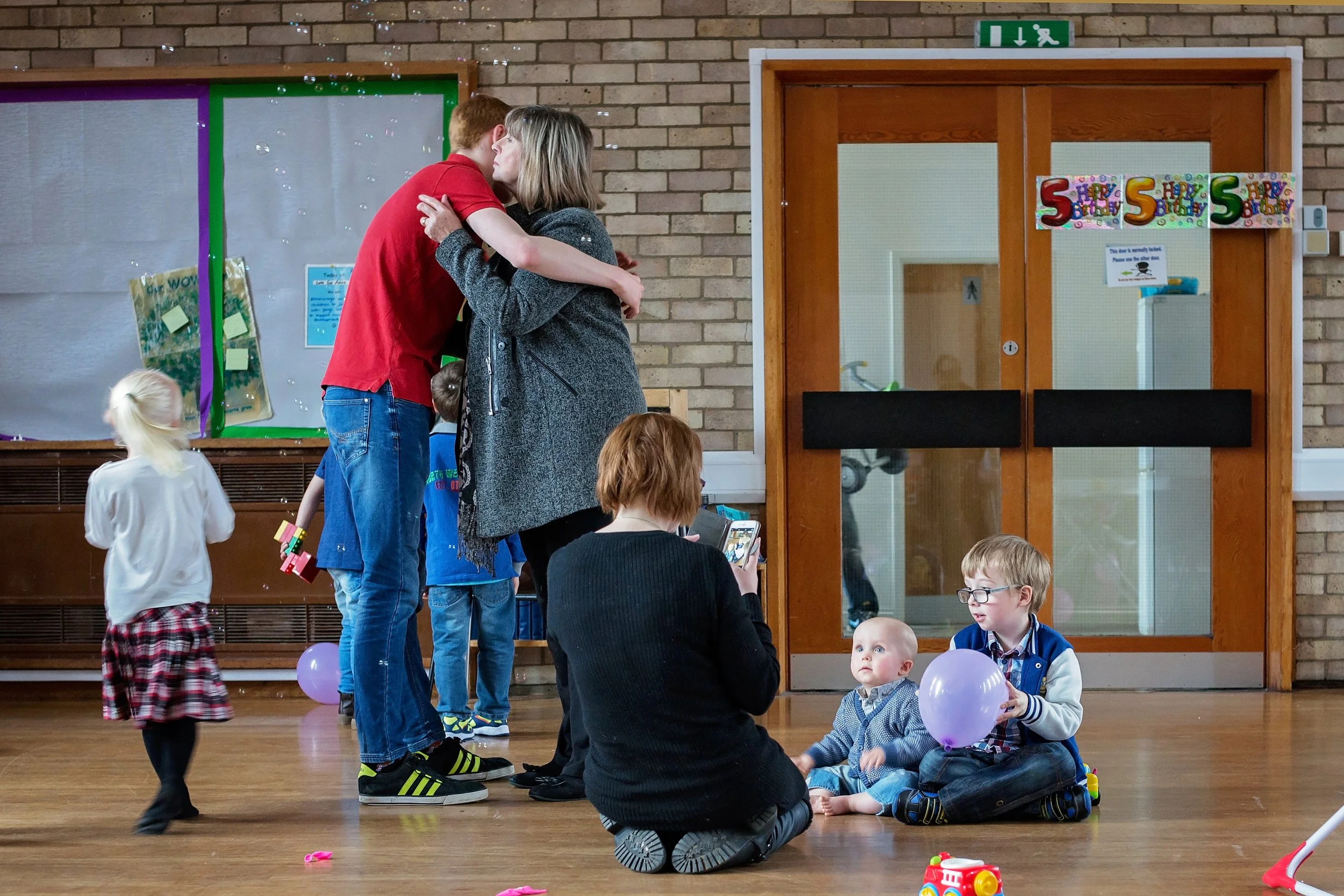 A group of children and adults at a birthday party in a decorated room. Two children sitting on the floor, one with a balloon, others standing or kneeling nearby. Two adults, one woman taking a photo, and a man kissing a woman.