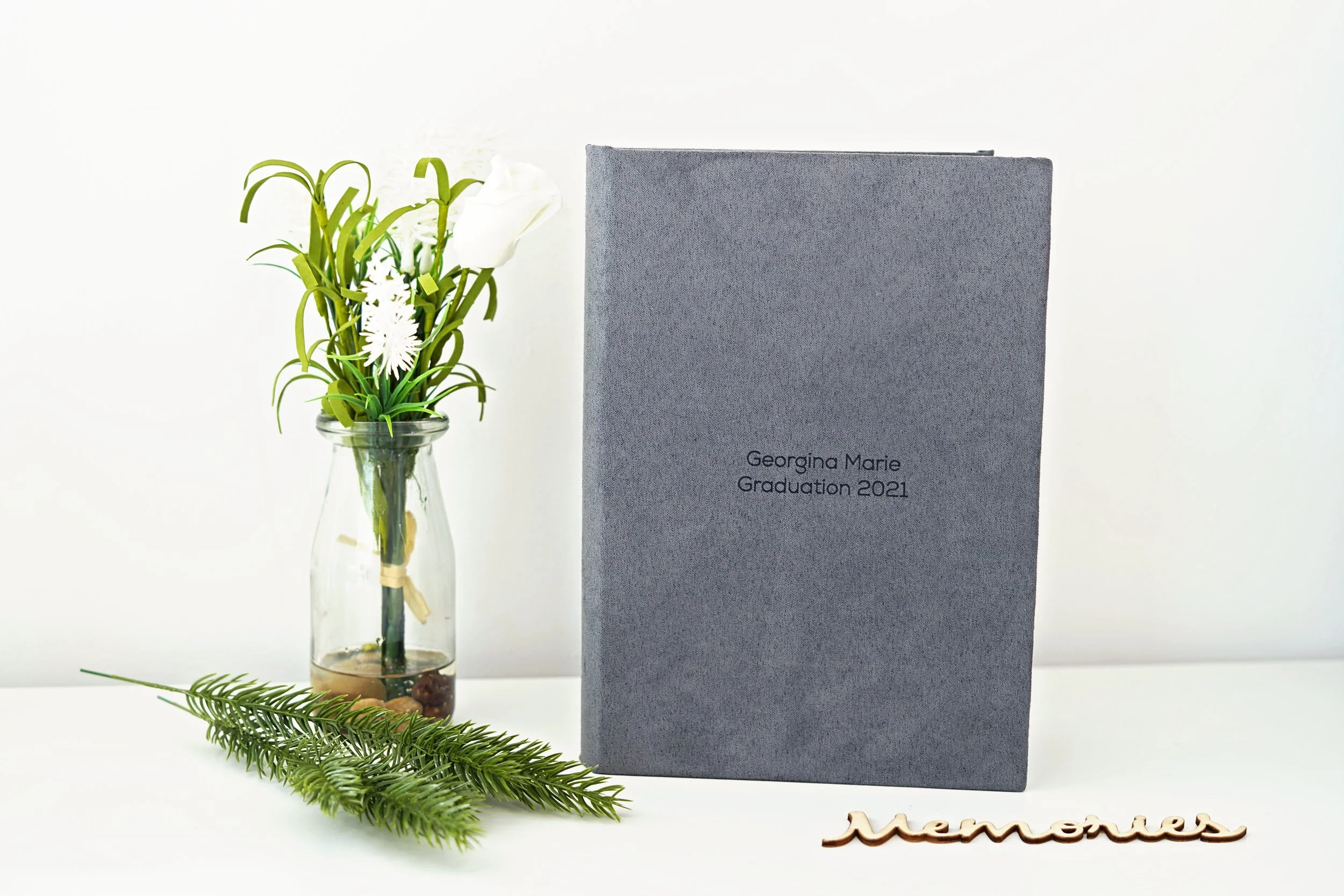 A gray graduation photo album with the inscription 'Georgina Marie Graduation 2021' placed on a white surface next to a glass vase with white flowers and greenery, and a sprig of pine, with the word 'Memories' spelled out in wooden letters.
