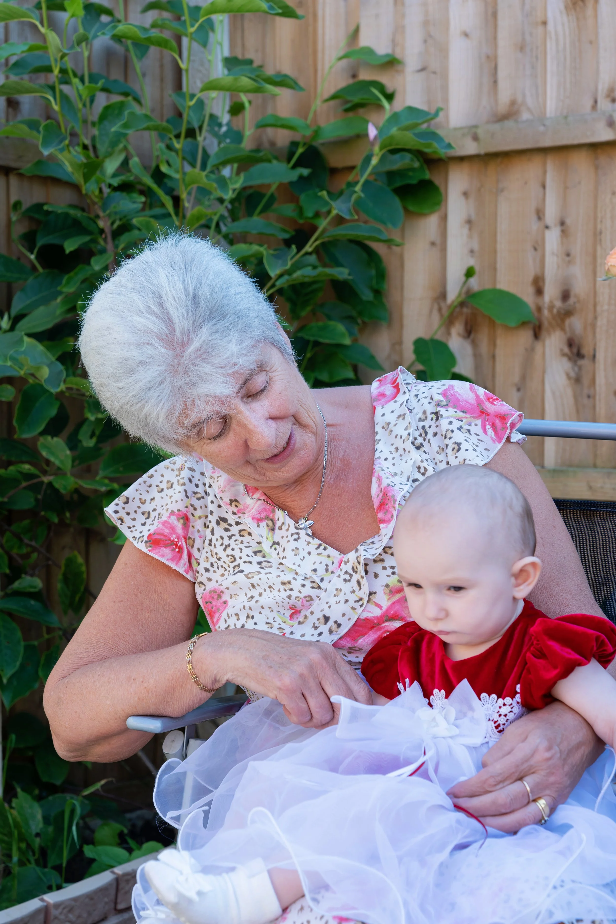 An elderly woman with white hair holding a young child in a red dress outside in a garden with a wooden fence and green plants.