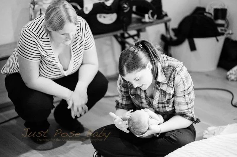 A woman holding a newborn baby while a second woman kneels beside her observing in a room with bags and a laundry basket in the background.