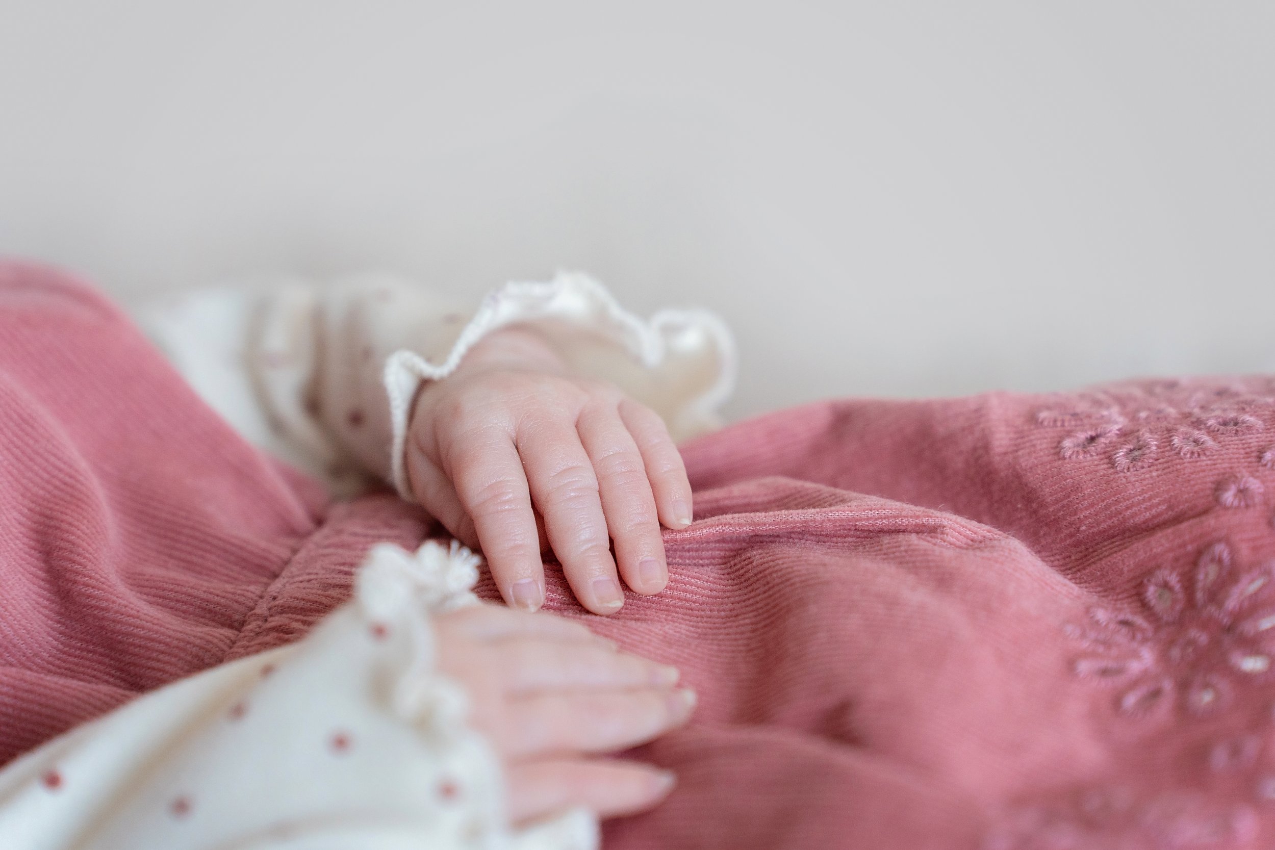 Close-up of a baby's hand resting on pink fabric with embroidery, wearing a cream-colored sleeve with ruffled edges.
