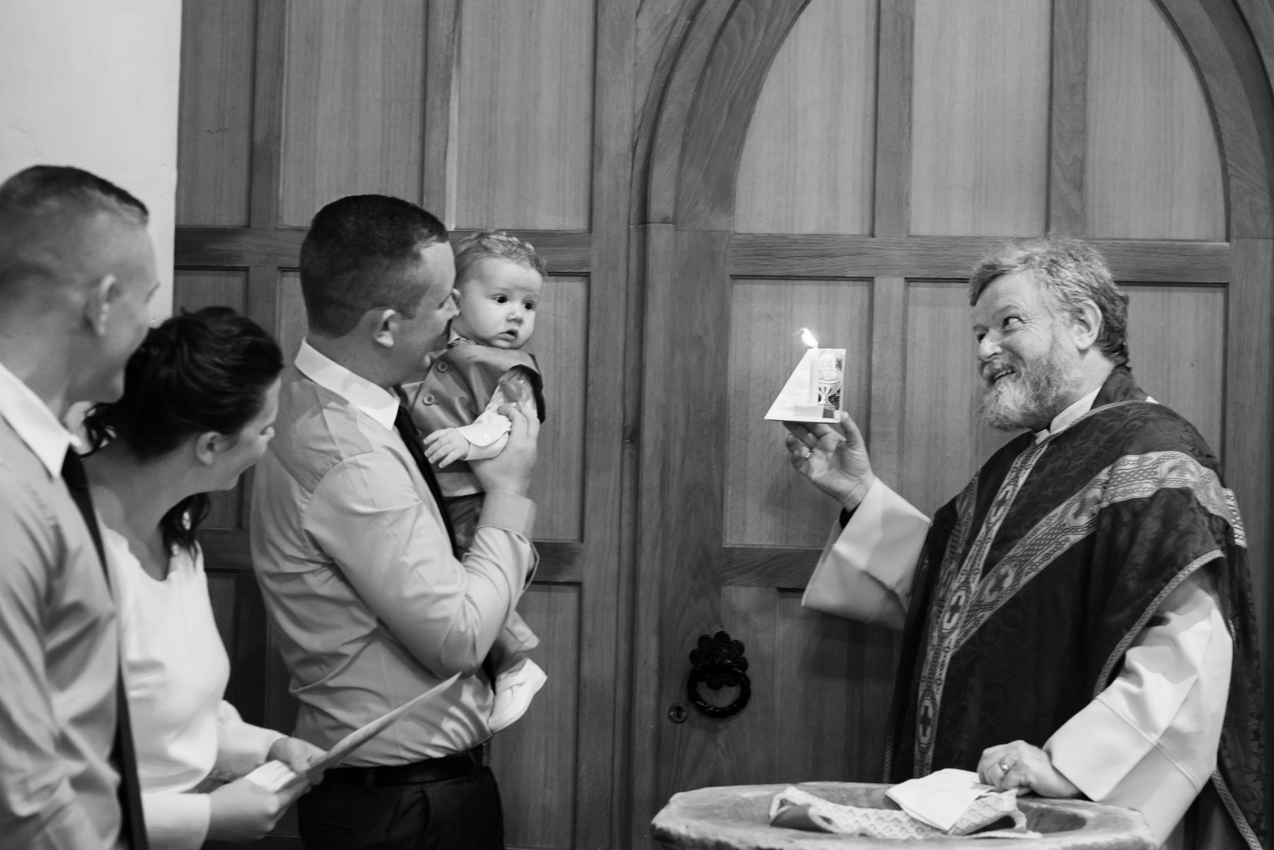 A priest conducts a baptism ceremony, holding a lit candle and a small booklet, while a family watched attentively, with a young child held by an adult. All are dressed in formal attire, inside a room with wooden paneled walls.
