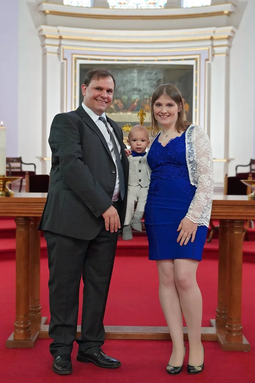 Family of three, man, woman, and baby, standing inside a church with red carpet, wooden altar, and religious artwork in the background. The man wears a dark suit, the woman wears a blue dress with lace sleeves, and the baby is dressed in a suit, sitting on the woman's arm.