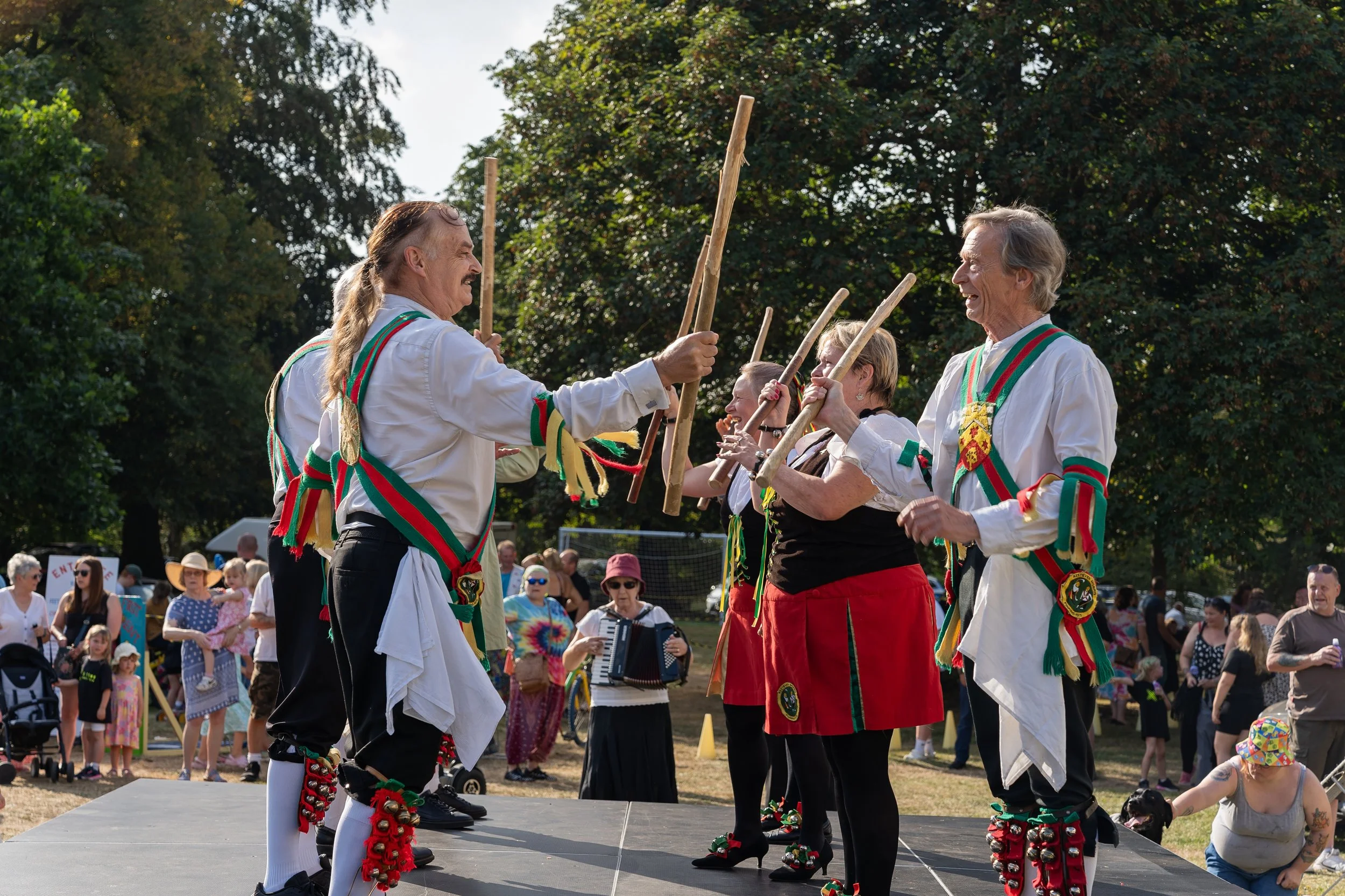 A group of people dressed in traditional folk costumes performing a dance with sticks at an outdoor festival. There are onlookers in casual clothing watching from the background.
