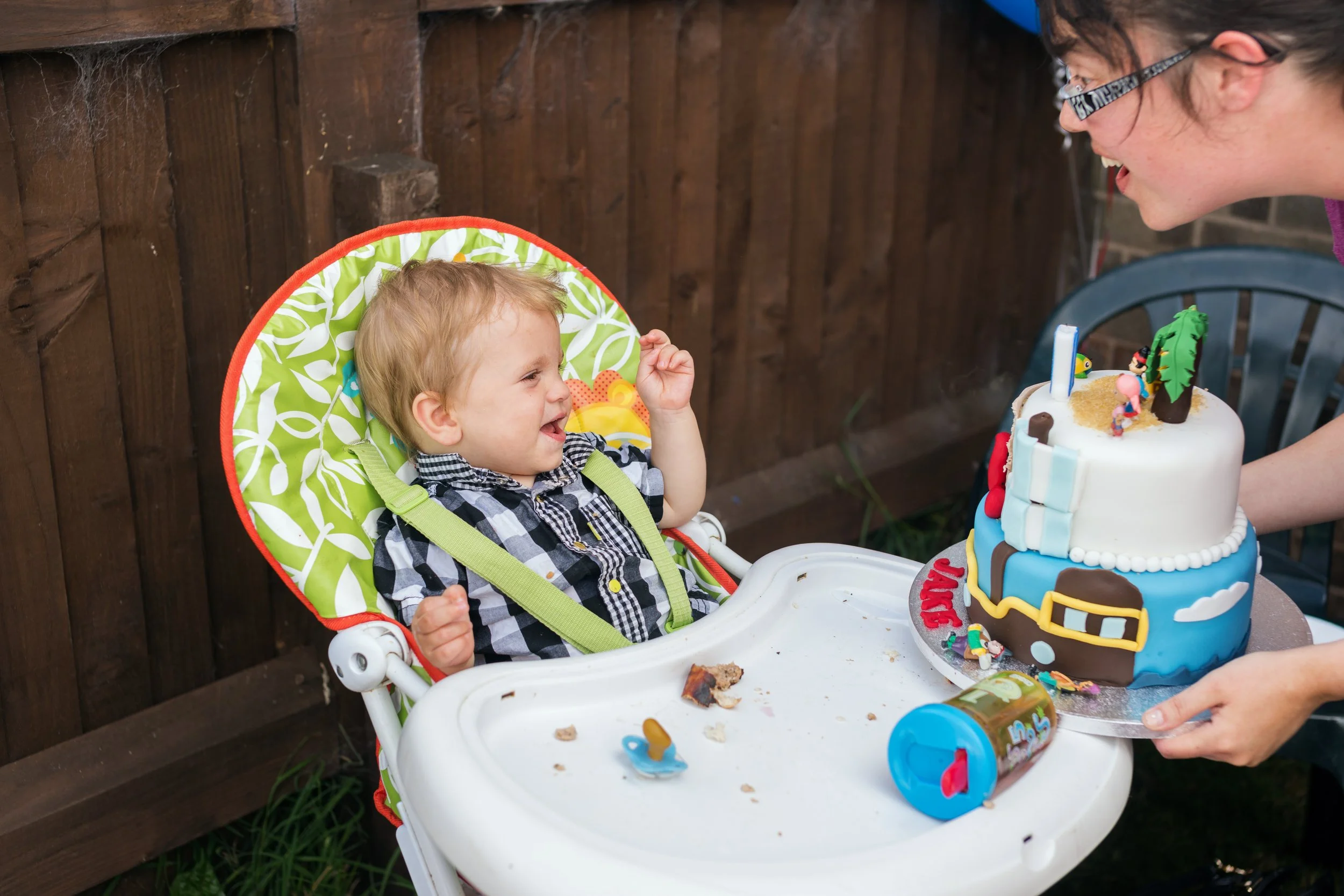 A young boy sitting in a high chair, smiling and celebrating his birthday with a birthday cake and a woman holding the cake towards him.