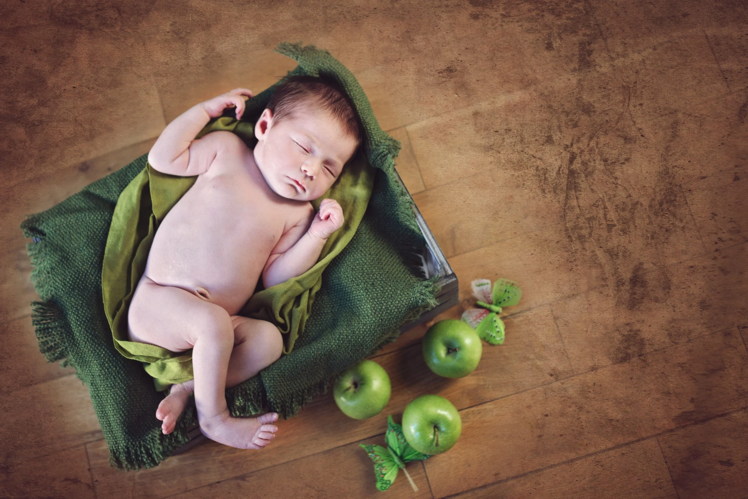 A sleeping newborn baby lying on a green blanket, surrounded by three green apples and two green butterfly decorations, on a wooden floor.