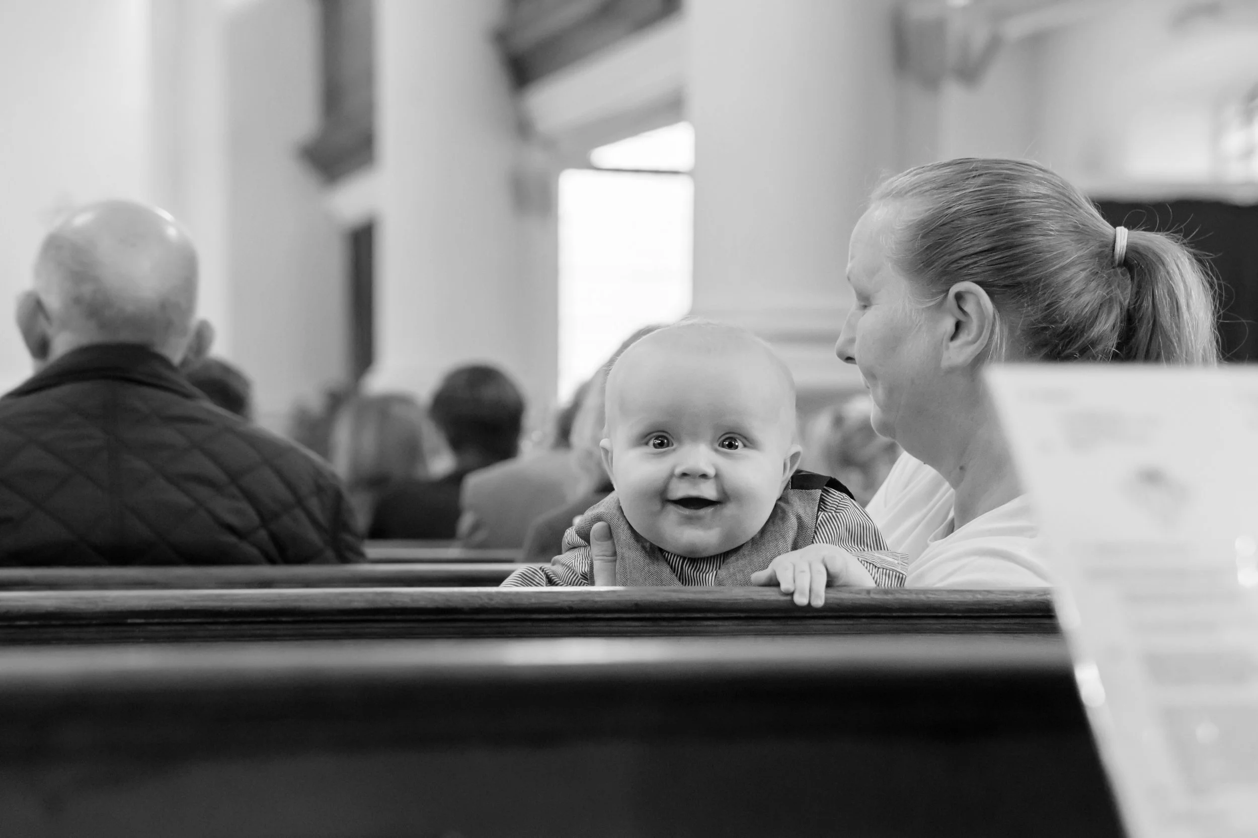 A baby leaning on a church pew, smiling, next to a woman, surrounded by other people in a church setting, in black and white.