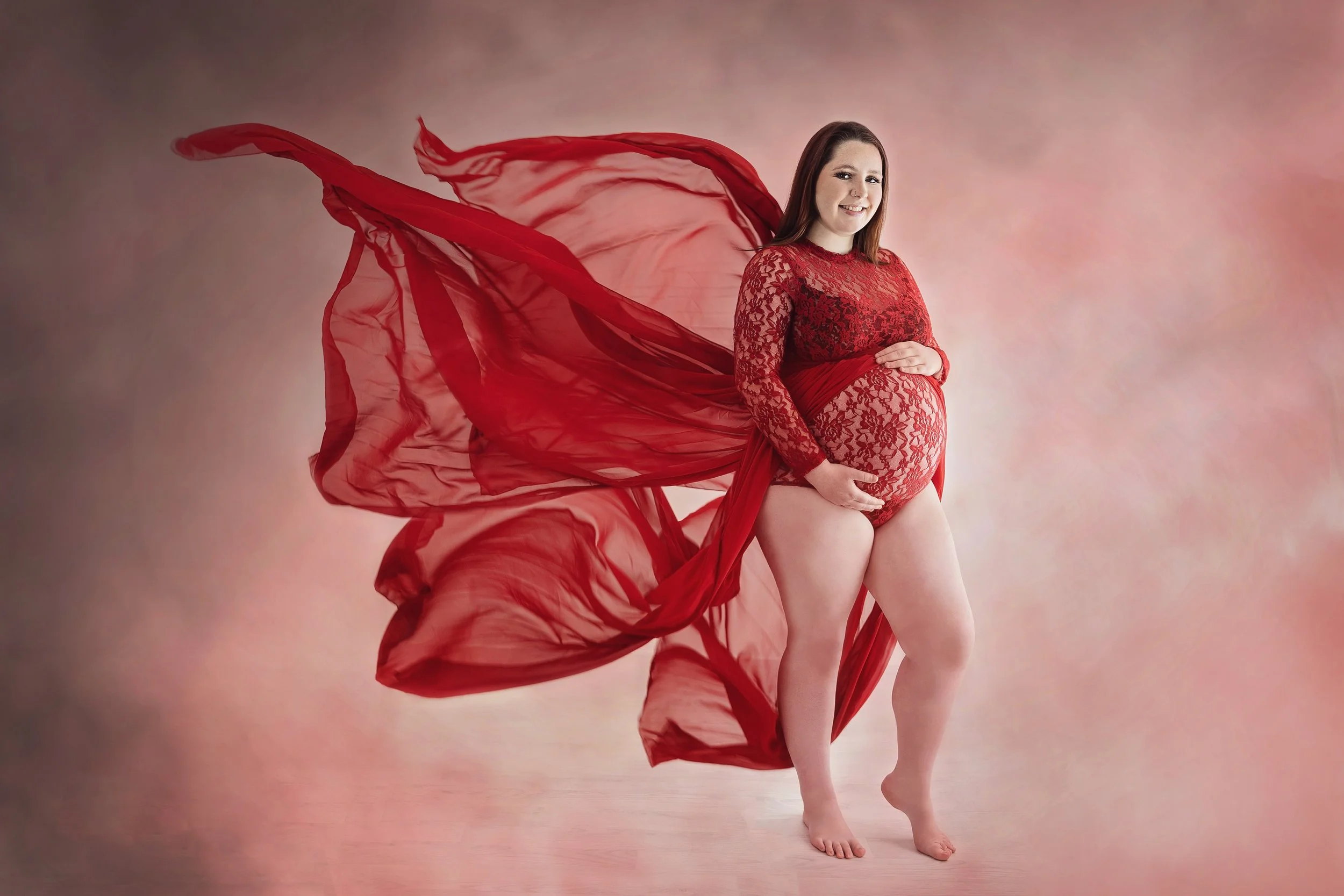 Pregnant woman in red lace outfit with flowing red fabric standing against a soft pink background.
