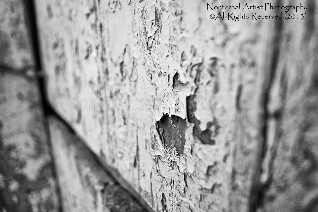 Close-up black and white photograph of peeling and chipped paint on a wooden surface.