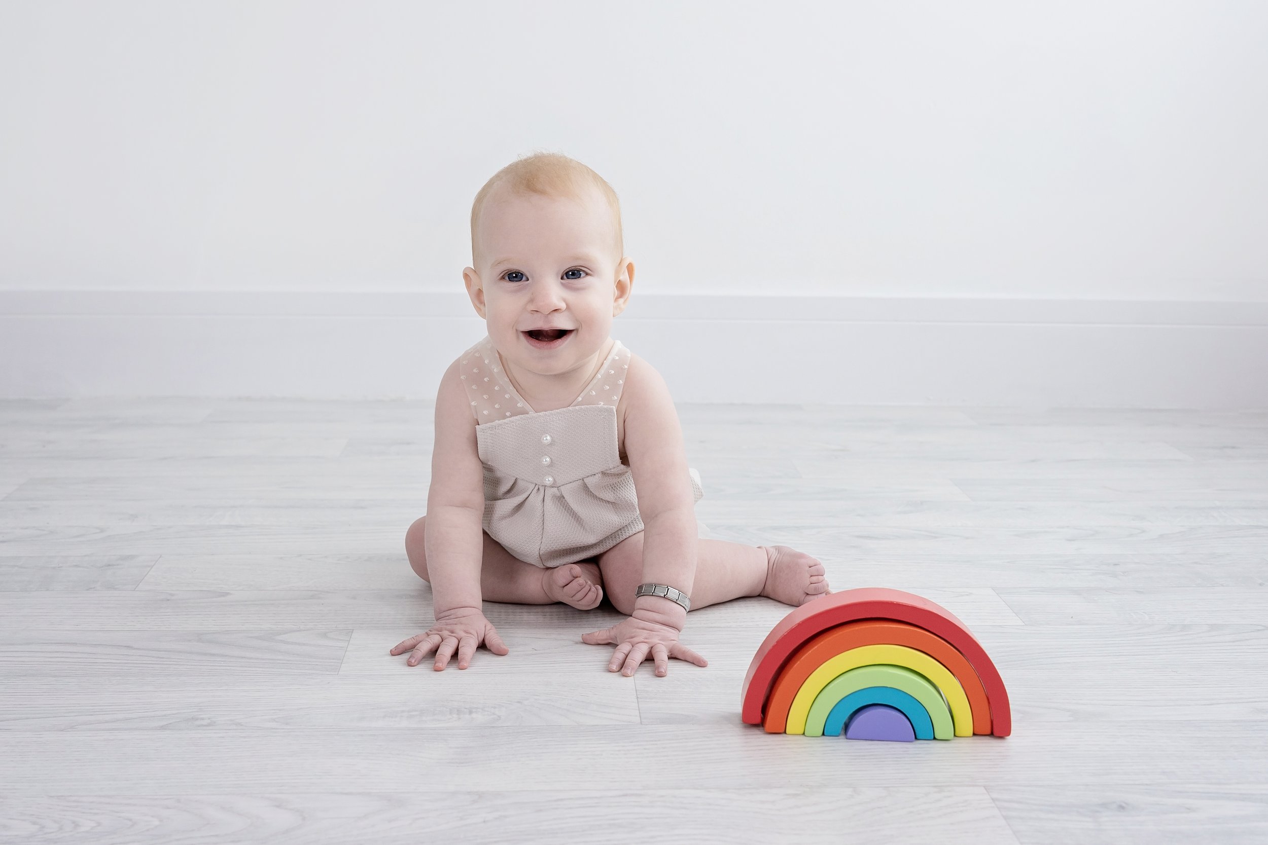 A smiling baby with light skin and blonde hair sitting on a white wooden floor next to a colorful rainbow toy.