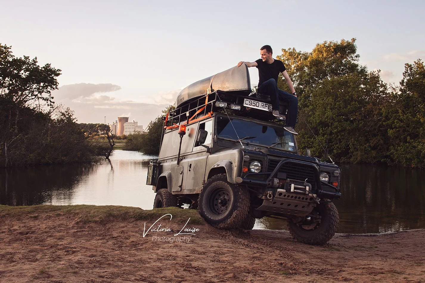 A man sitting on top of a black off-road vehicle with a roof rack, parked on sandy ground beside a river, surrounded by trees, during sunset or late afternoon.