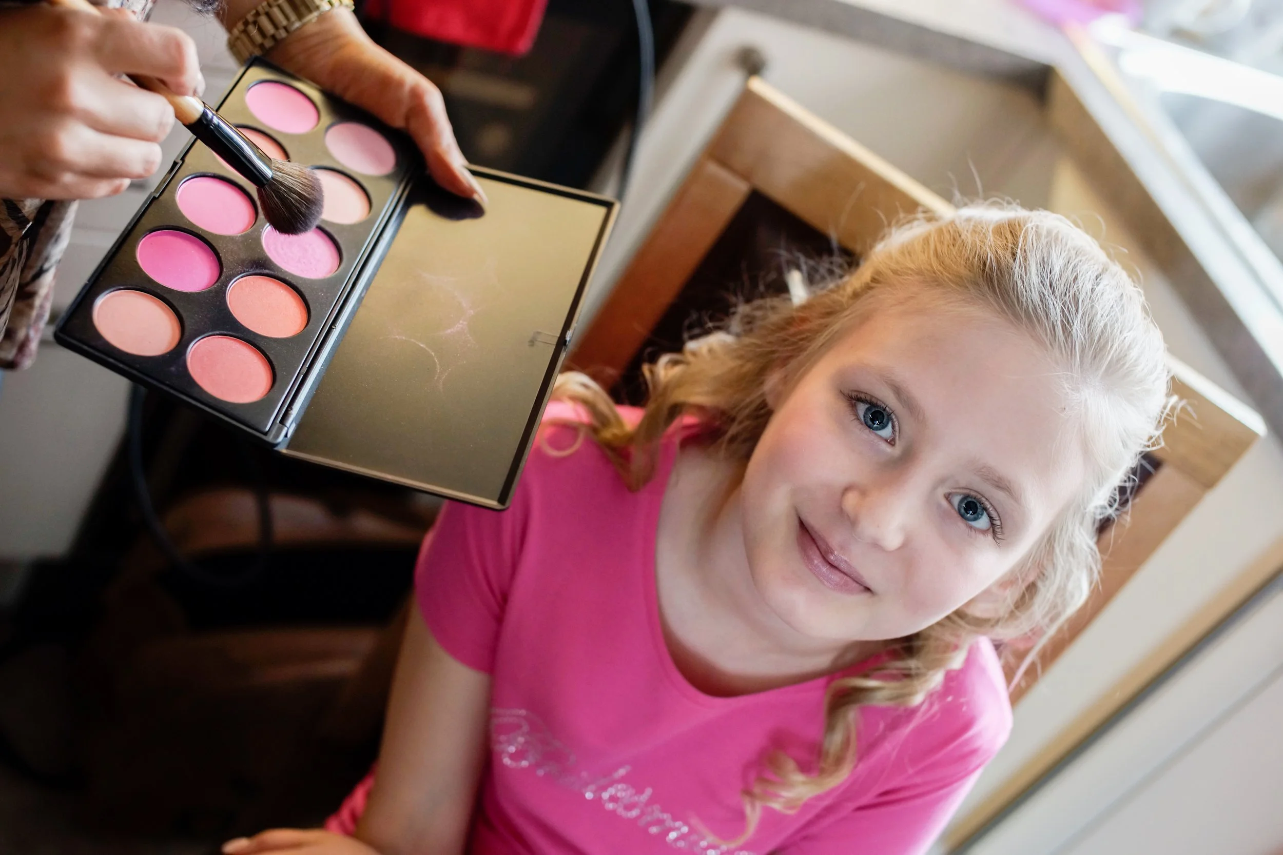 Young bridesmaid is getting her make up done, she is looking directly up at the camera, smiling. The make up pallet can be seen by the side of her.