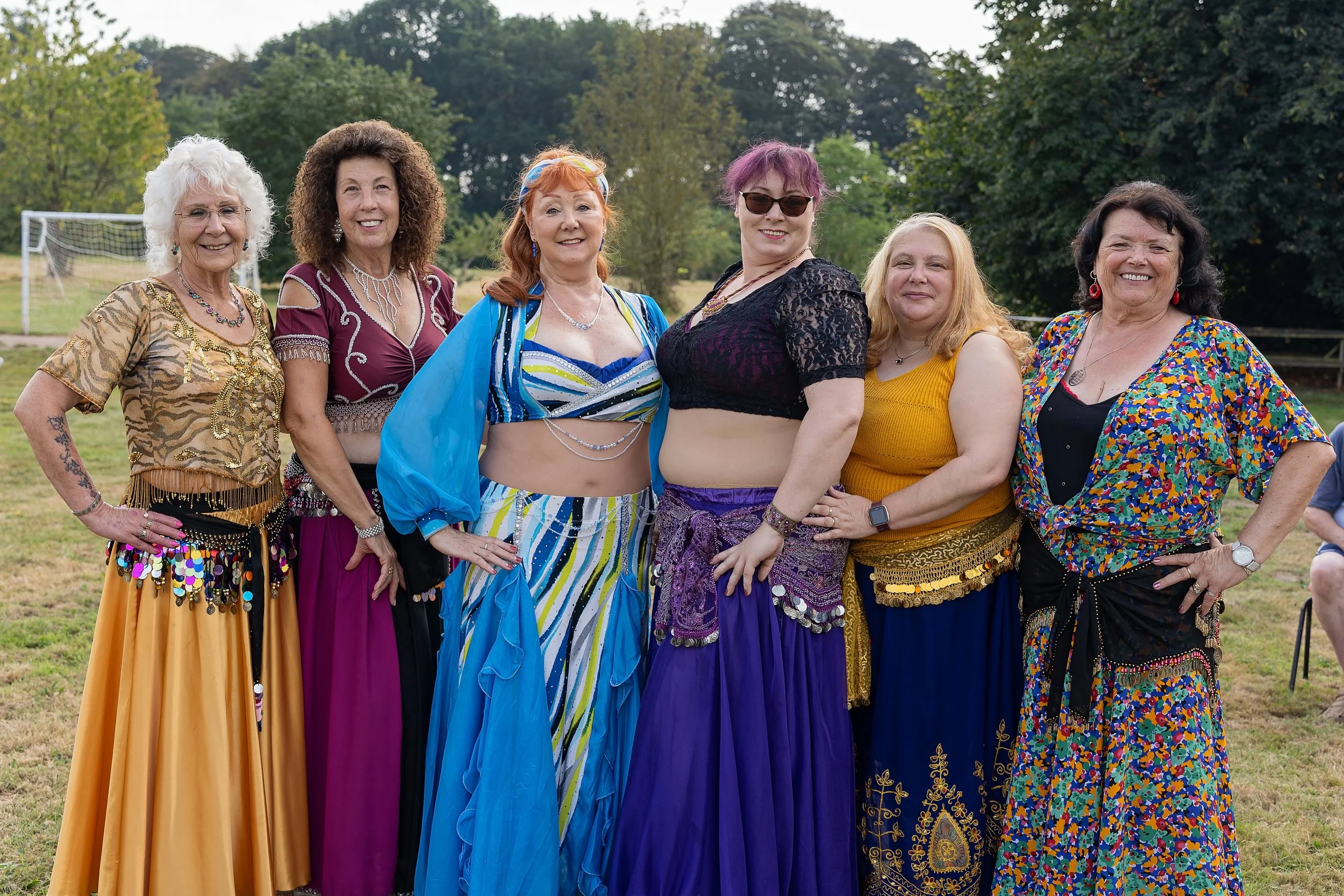Group of seven women dressed in colorful bohemian and belly dance-inspired outfits standing outdoors in a field with trees in the background.